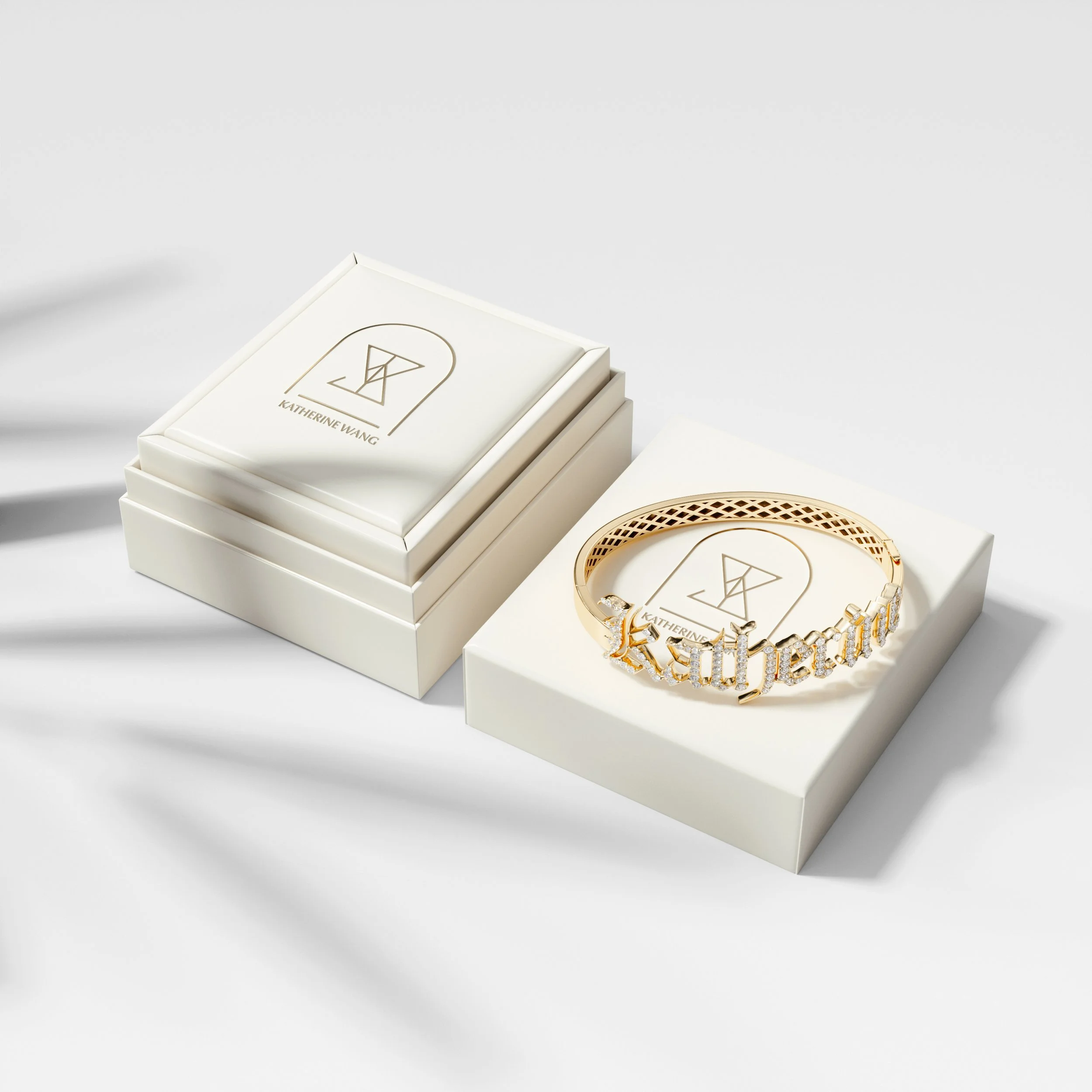 A gold bracelet with the name 'Katherine Wang' spelled out in diamond-studded letters, placed in a white box with a matching lid. The lid features a logo with a minimalist geometric design and the name 'Katherine Wang' underneath.