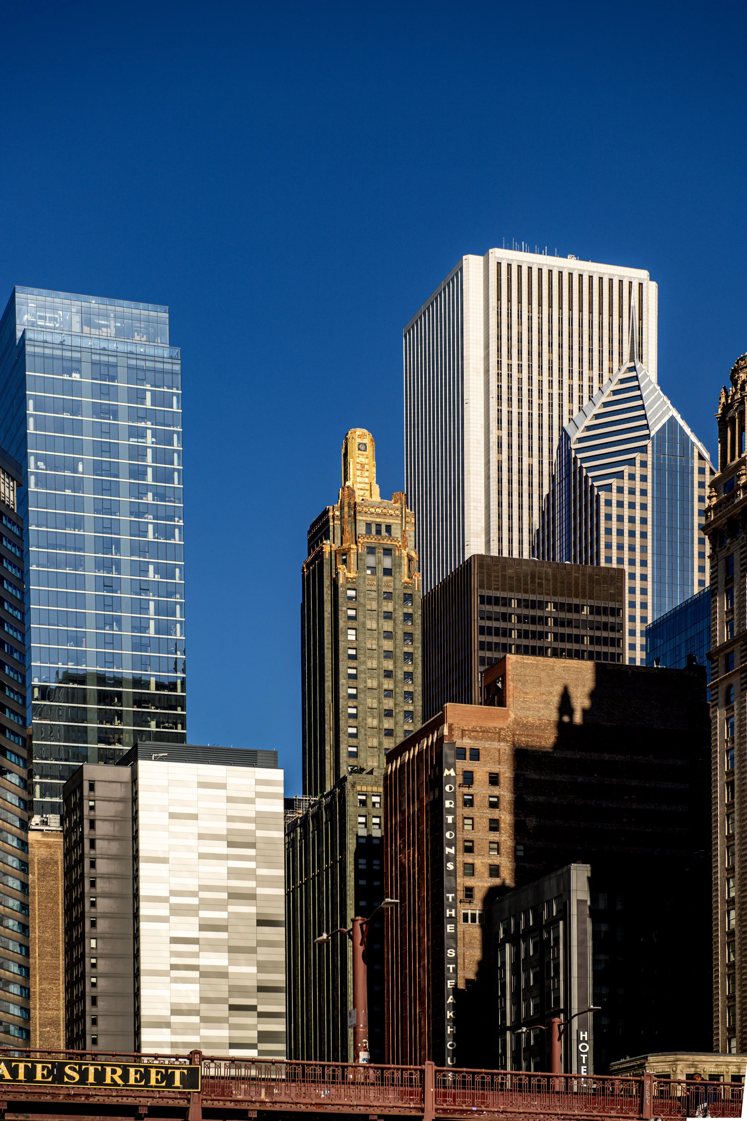 Skyline view of downtown buildings, including a tall glass skyscraper on the left, a gold-topped art deco building in the center, and other modern high-rises against a clear blue sky.