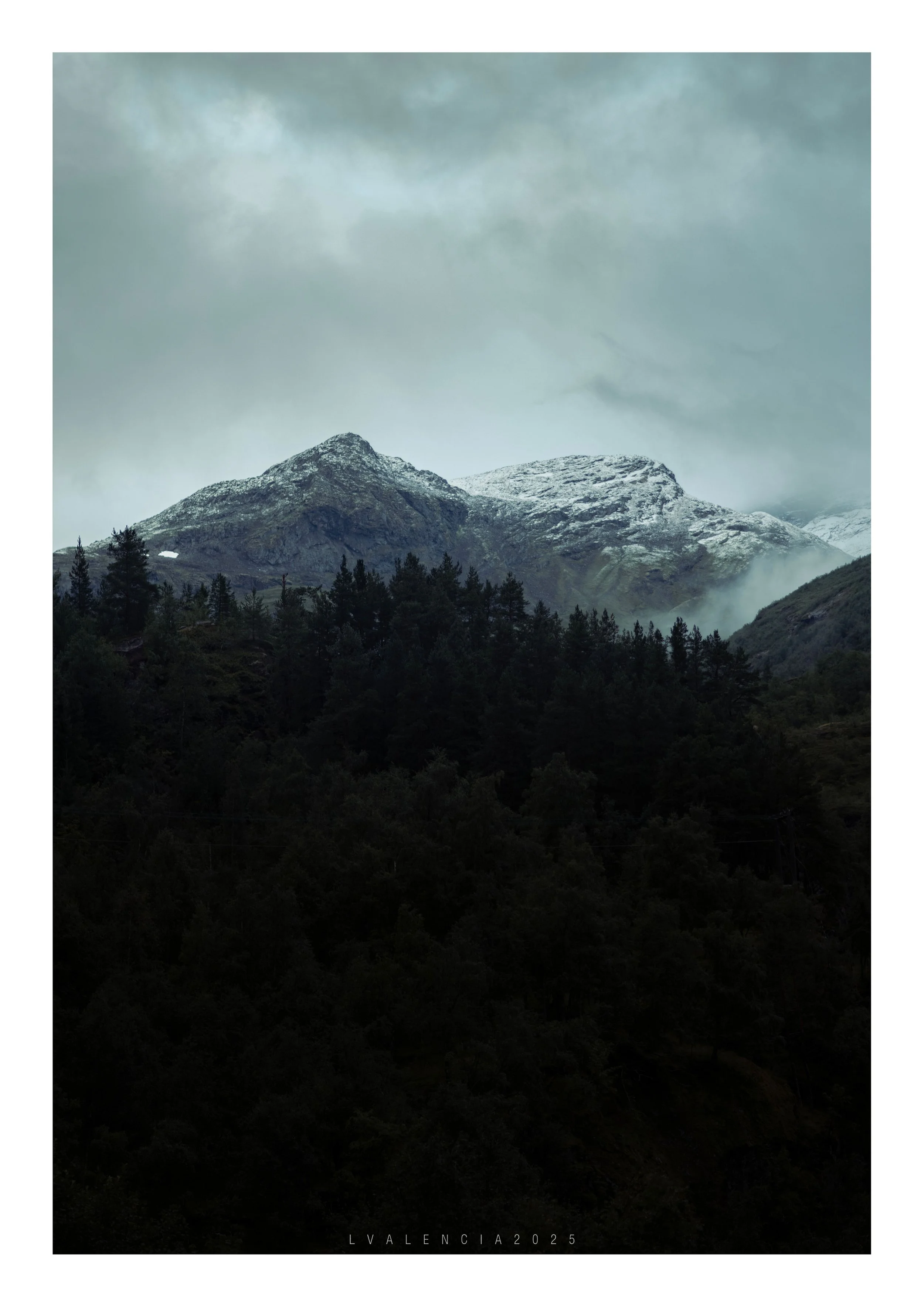 Mountain landscape with snow-capped peaks, forested slopes, and cloudy sky.