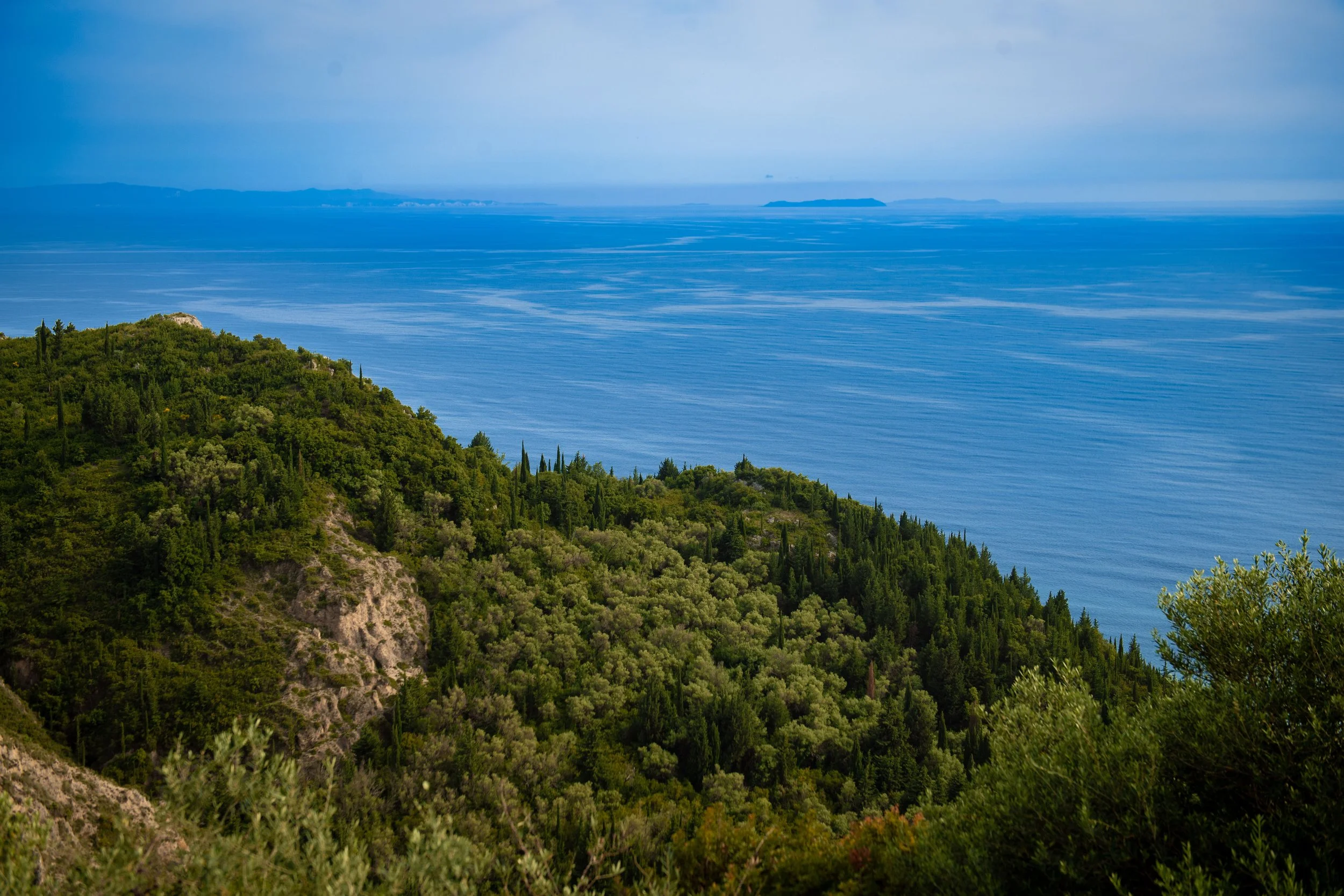 A scenic view of lush green hills and trees leading down to the ocean, with small islands and a cloudy sky in the distance.
