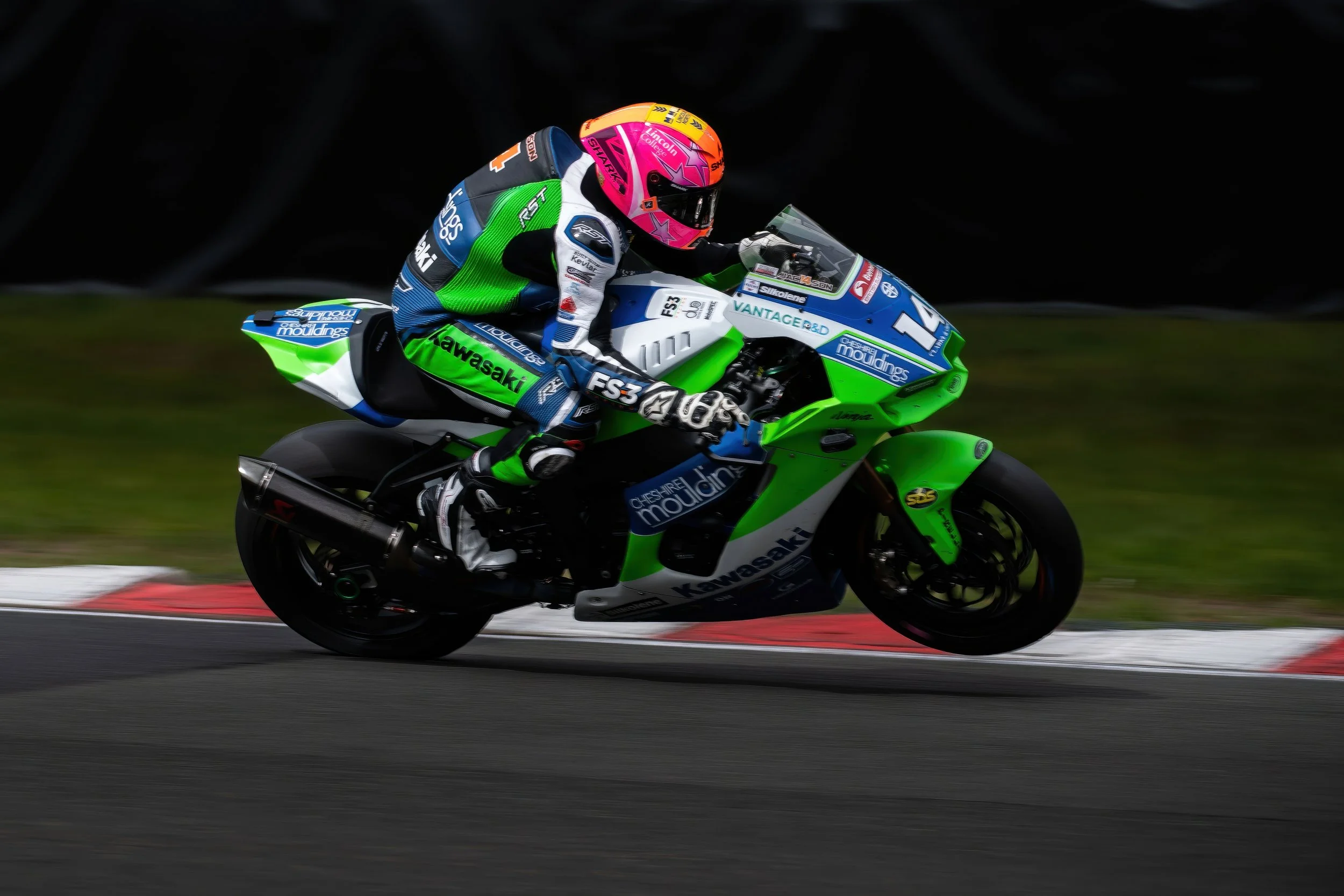 A motorcycle rider in racing gear leaning into a turn on a race track, riding a green, black, and blue Kawasaki motorcycle with various sponsor logos. The rider is wearing a pink helmet and a colorful suit.