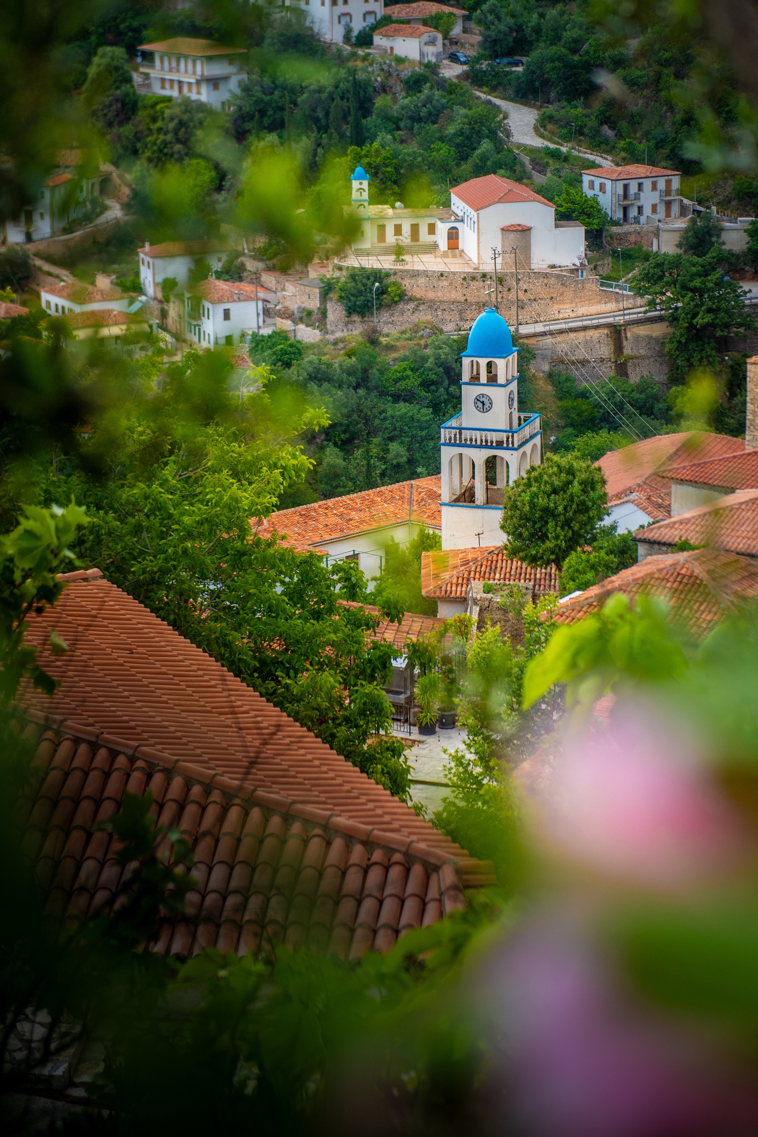 A scenic view of a hillside village with white buildings, terracotta rooftops, and a prominent white and blue church tower with a clock, surrounded by green trees.