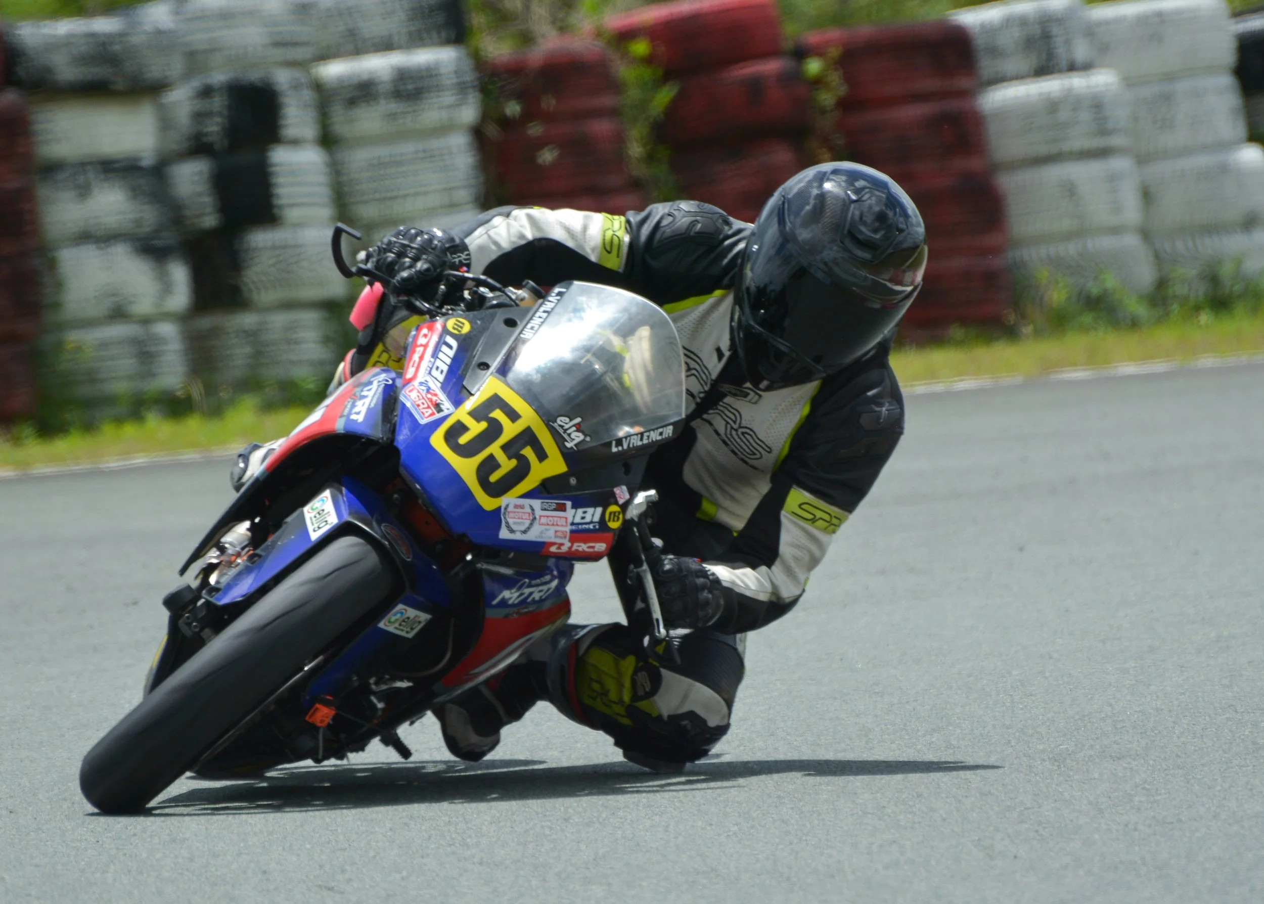 Motorcycle racer in black and white gear with yellow accents leaning into a turn on a racetrack, with barriers in the background.