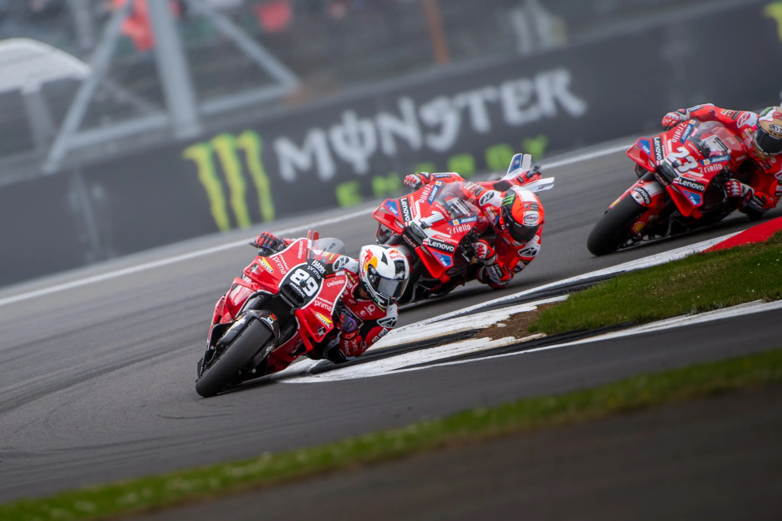 Three professional motorcycle racers in red racing suits leaning into a turn on a racetrack with a Monster Energy banner in the background.