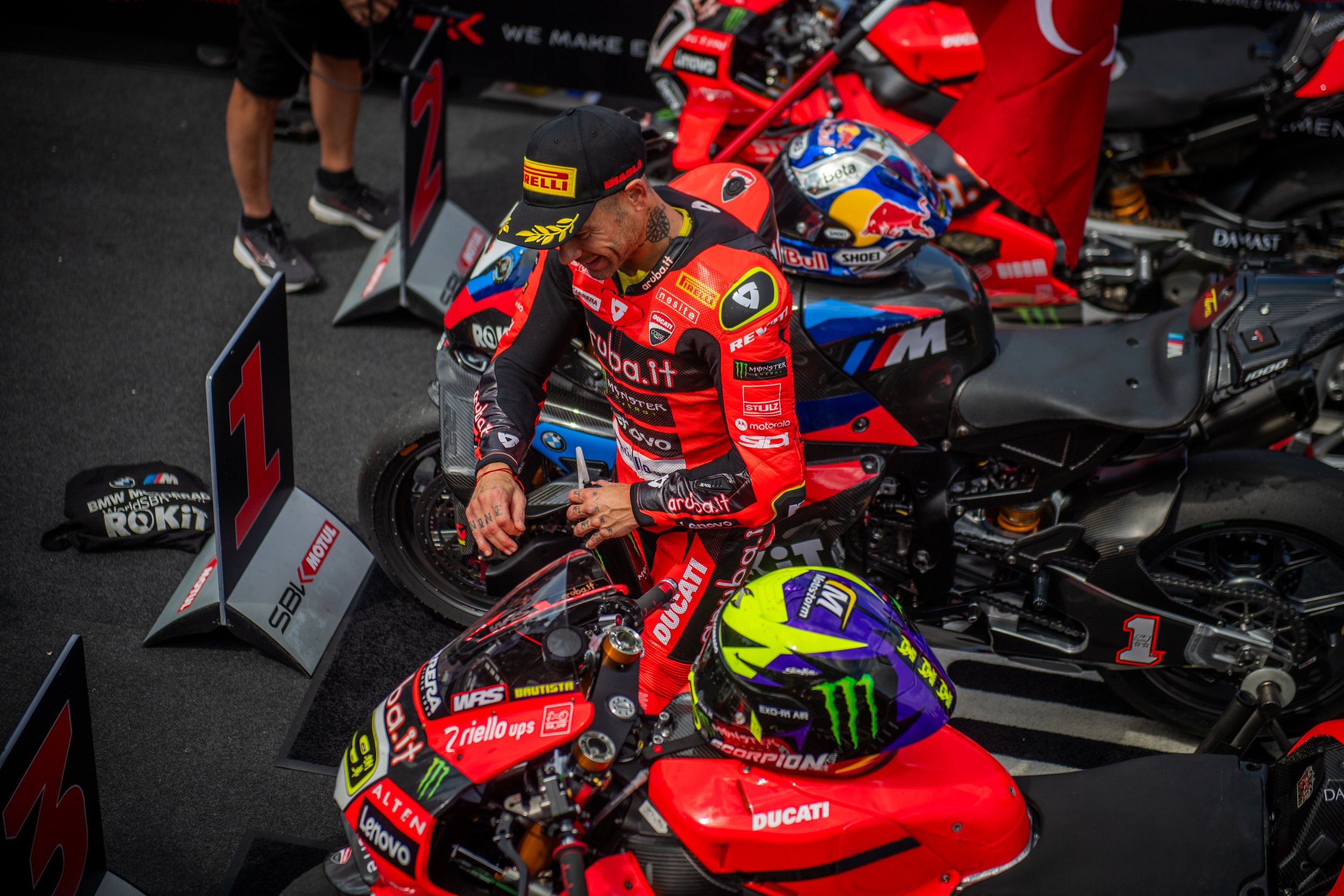 A motorcycle racer in red racing gear and a black cap signing an autograph next to his red Ducati motorcycle, with a blue and red motorcycle nearby in a paddock area.