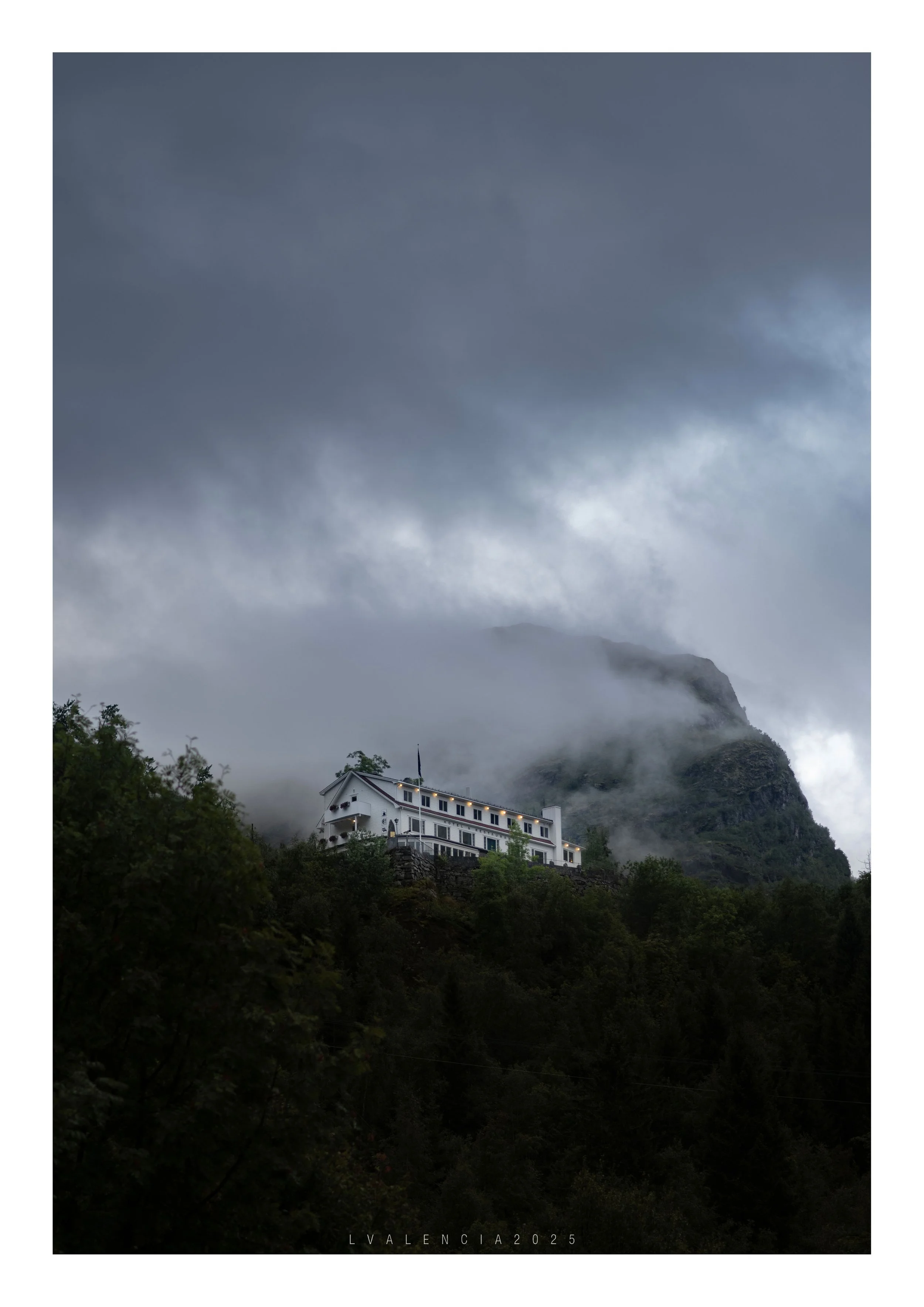 A large white house or hotel on a hillside with dense greenery, set against a background of misty mountains and dark stormy clouds.