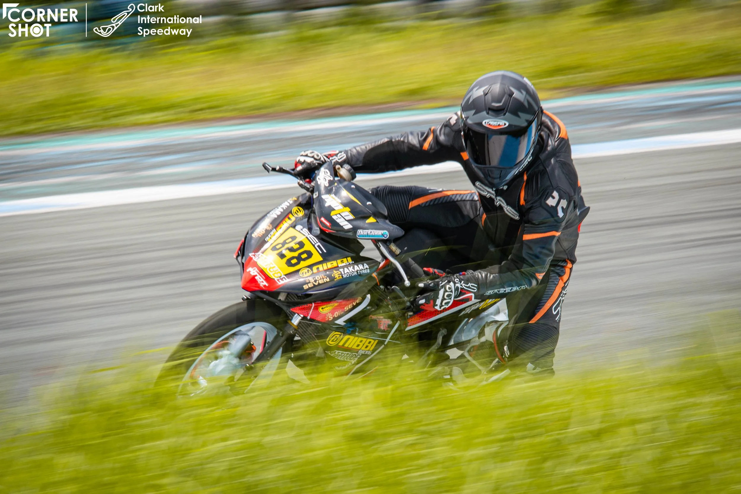 Motorcycle racer in black suit leaning into a turn on a racetrack, with grass and track markings visible in the background  rider name Lawrence Valencia