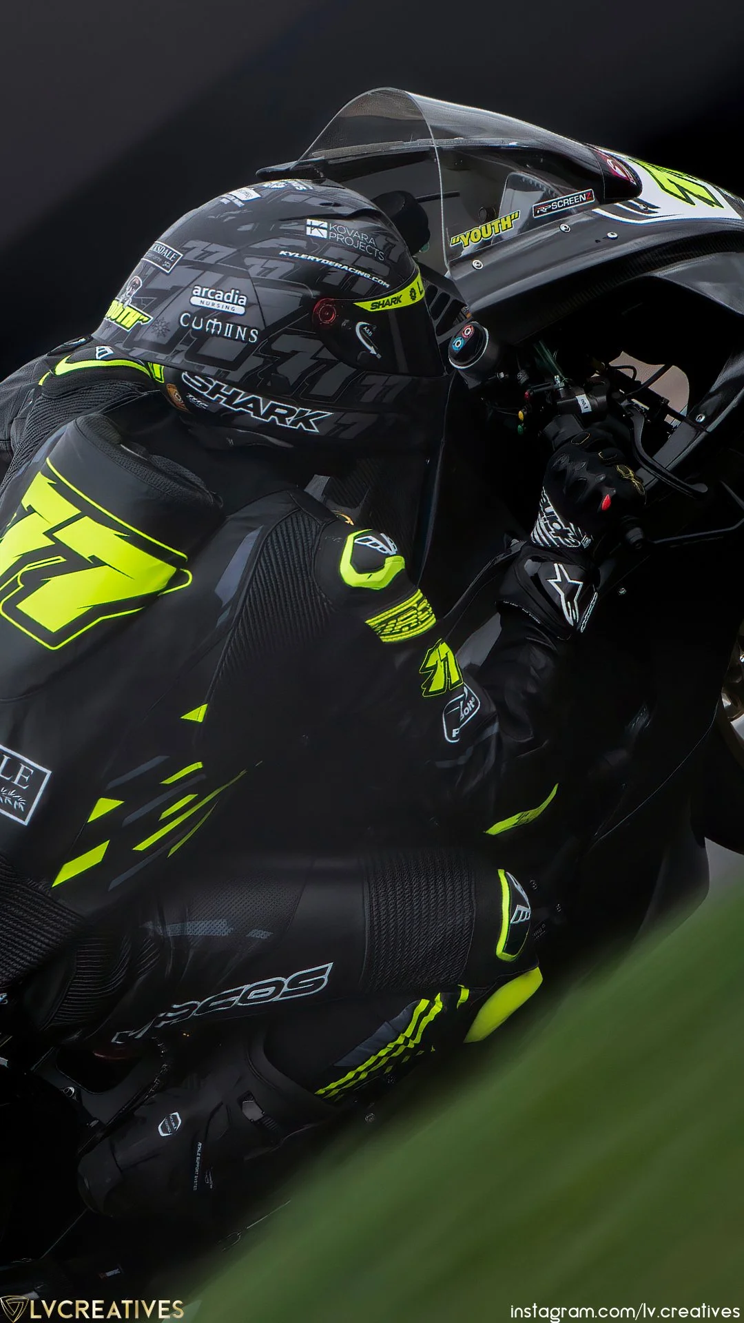 A race car driver wearing black and neon yellow racing gear and helmet, seated in a black race car, gripping the steering wheel.