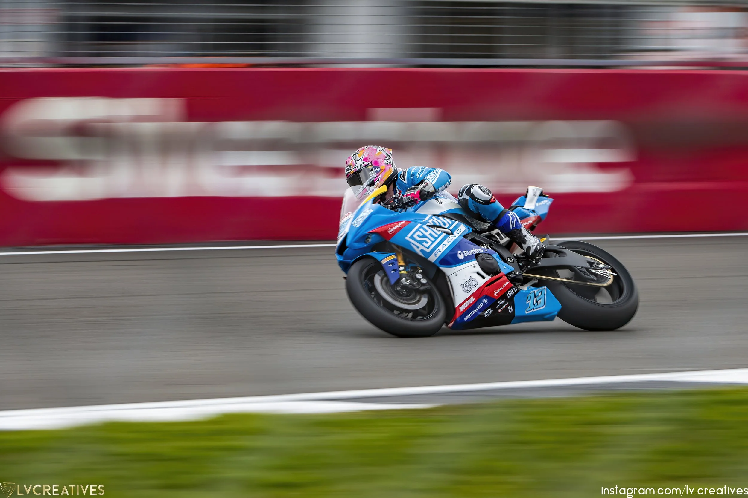 Motorcycle racer in blue racing suit leaning into a turn on a racetrack, with a red barrier and blurred background.
