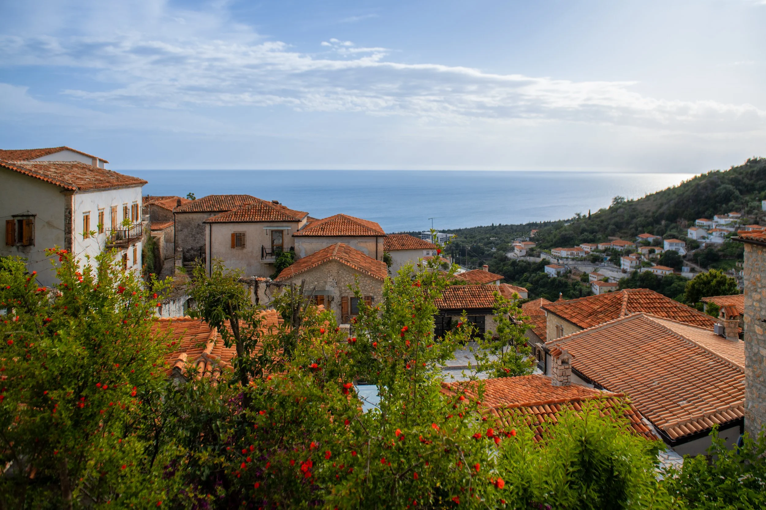 A scenic view of a Mediterranean village with terracotta rooftops, surrounded by lush greenery, overlooking the blue sea under a partly cloudy sky.