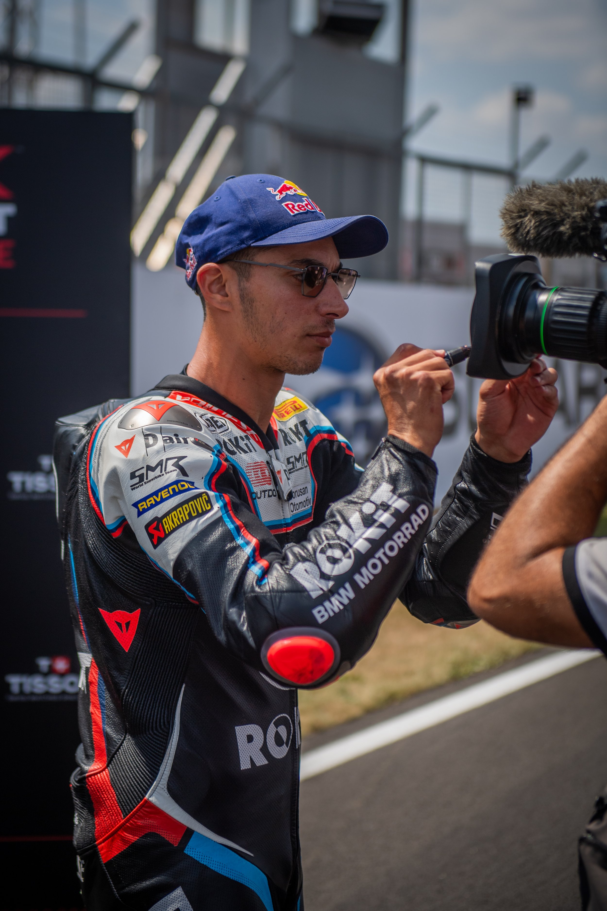 A man in a racing suit and sunglasses is at a racing event, signing an autograph, with a camera person capturing the moment.