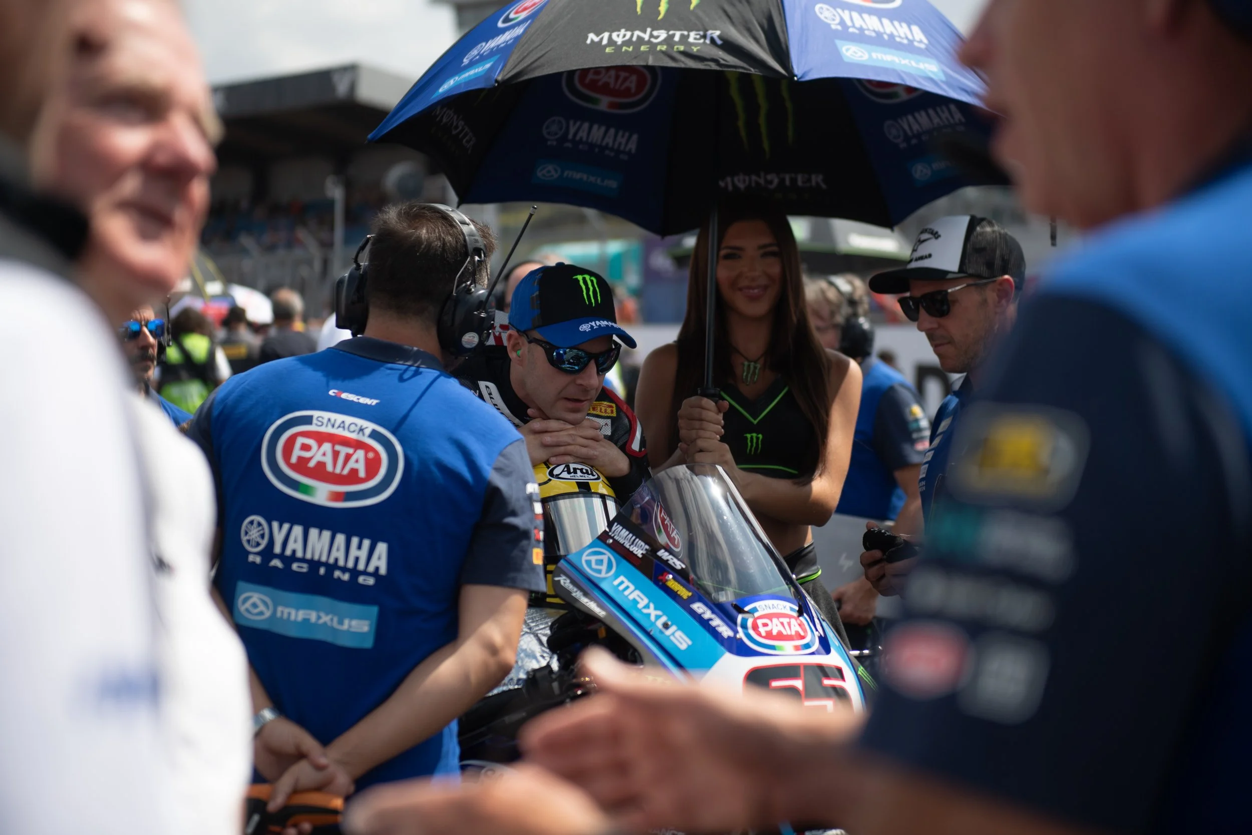 A race car driver in a racing suit sits on a motorcycle with a group of team members and a woman holding an umbrella around him, at a racing event. The team members wear blue uniforms with sponsor logos, and the woman and some team members wear sungl