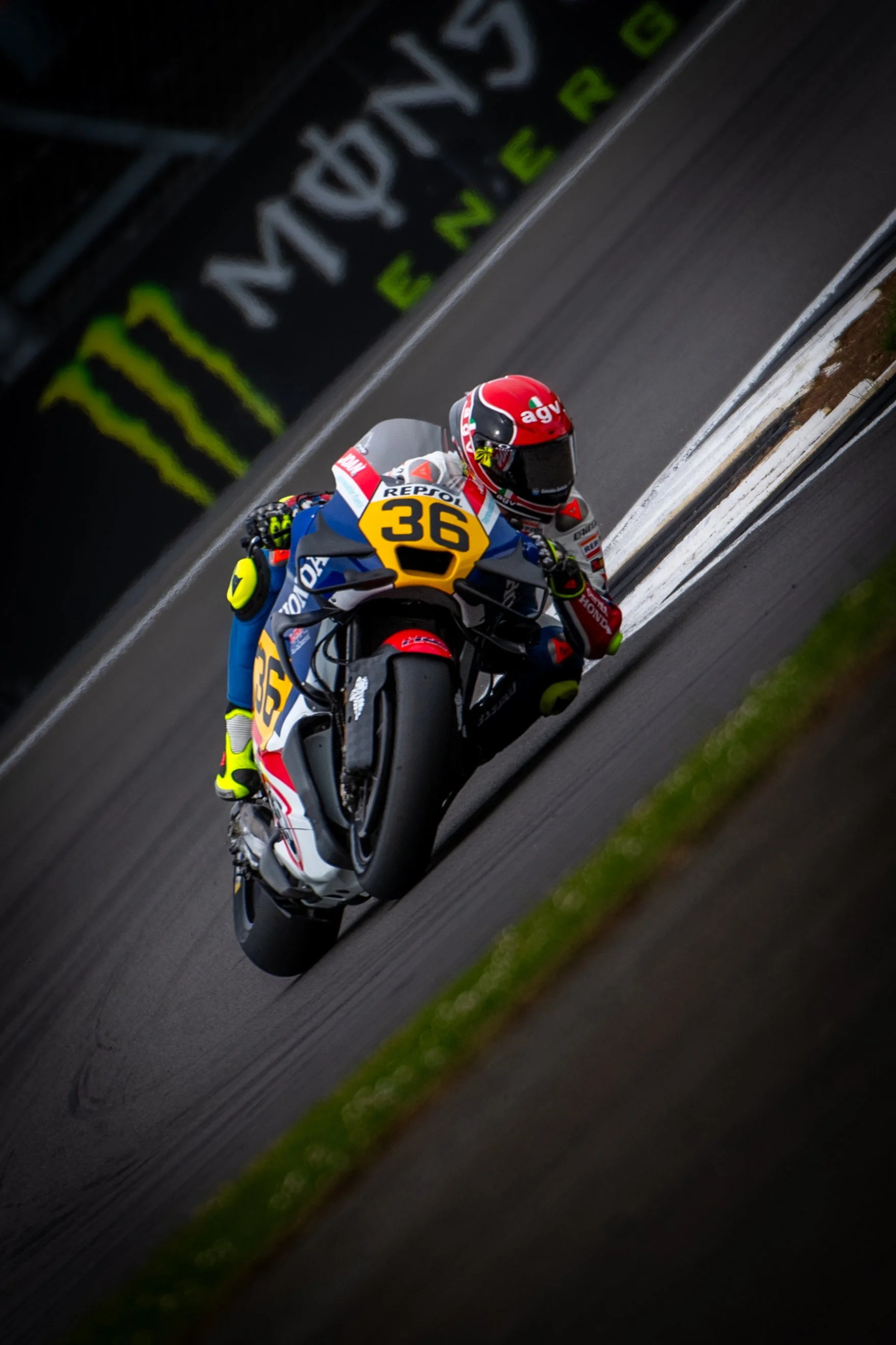 Motorcycle racer wearing red helmet, black visor, and colorful racing suit, riding a number 36 Repsol Honda motorcycle on a race track with advertising banners in the background.