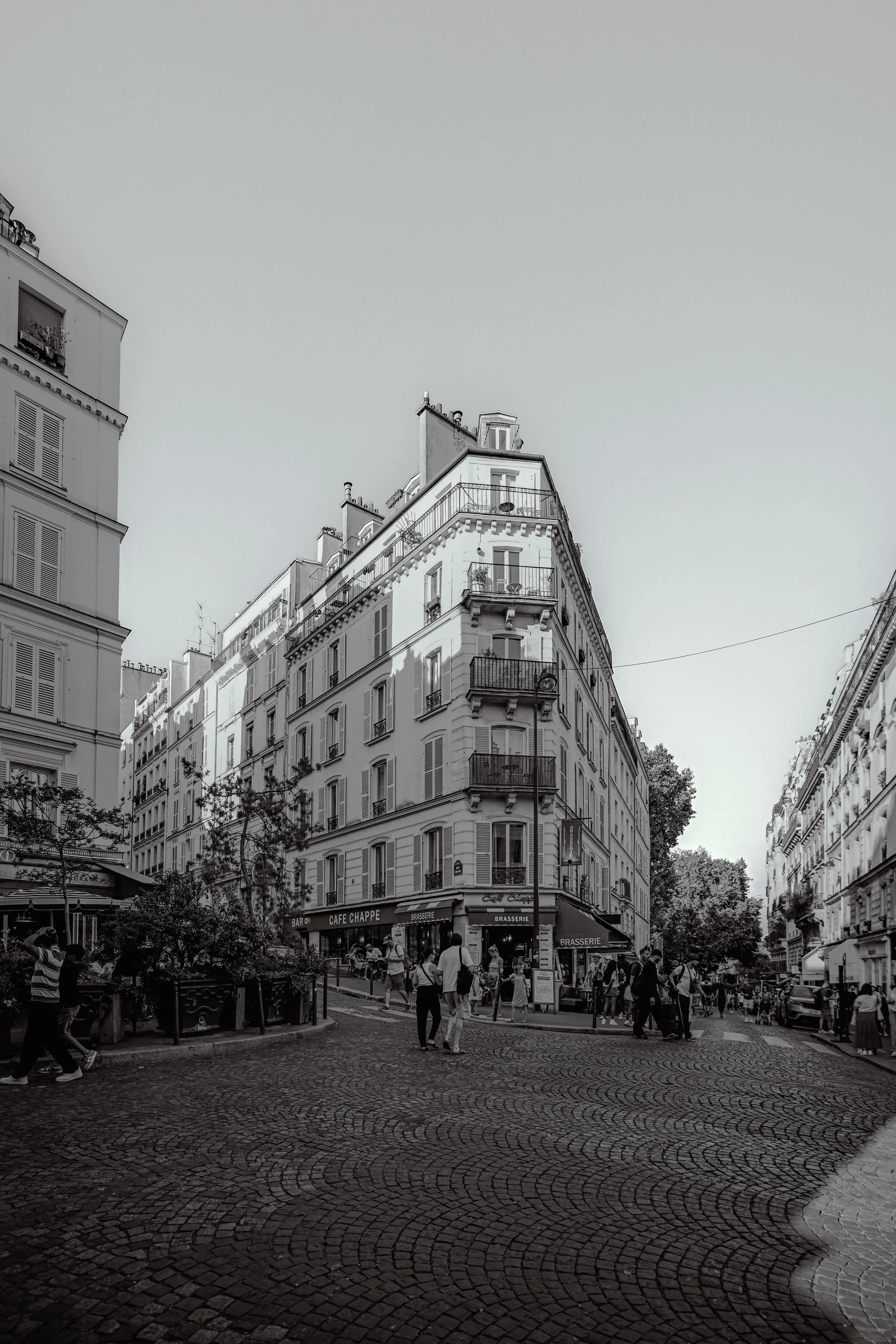 A black and white photo of a Parisian street corner with multi-story buildings, shops, and people walking on the cobblestone street.
