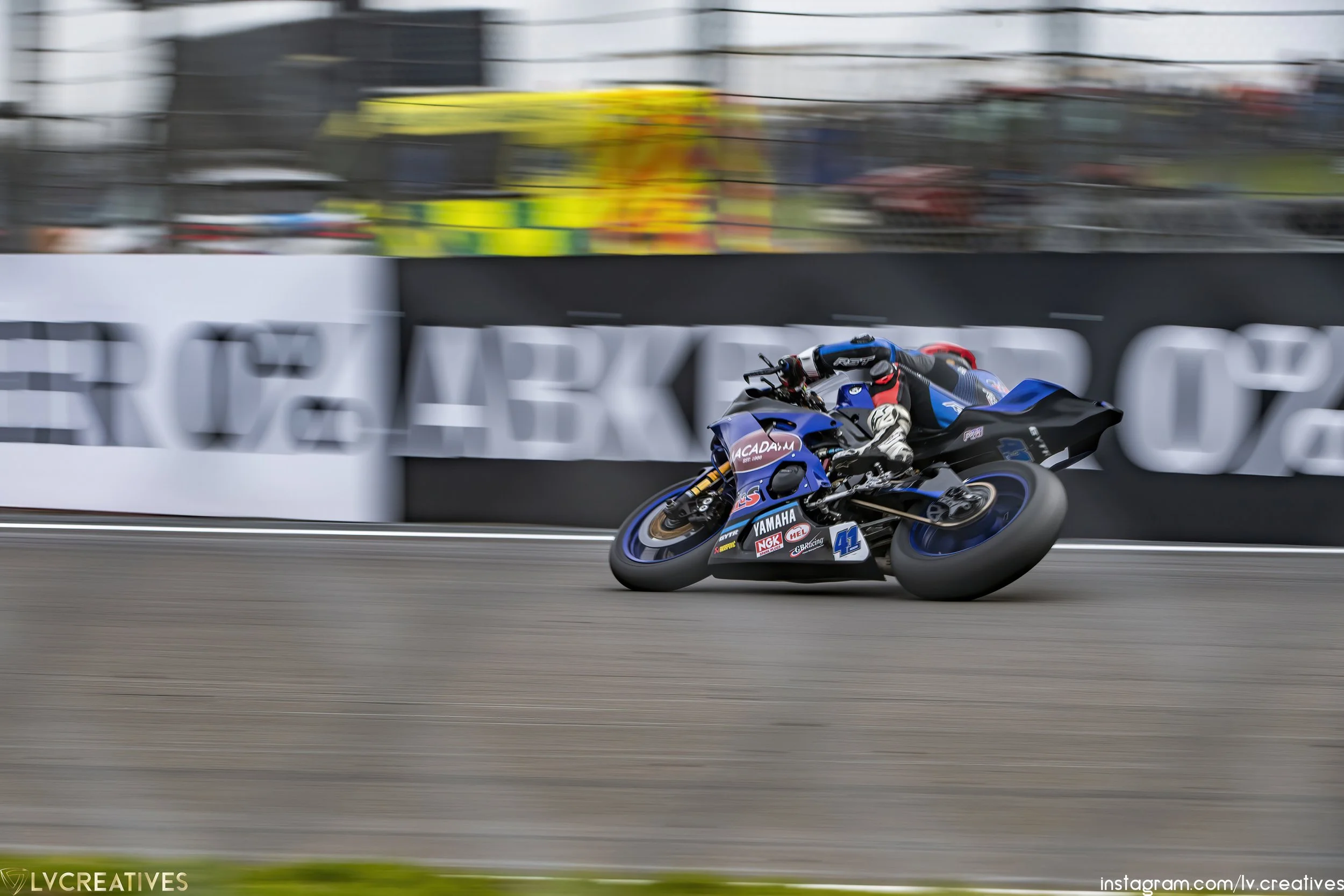 A motorcycle racer leaning into a turn on a racetrack. The bike is blue with various sponsor logos. The background shows blurred spectators and barriers.