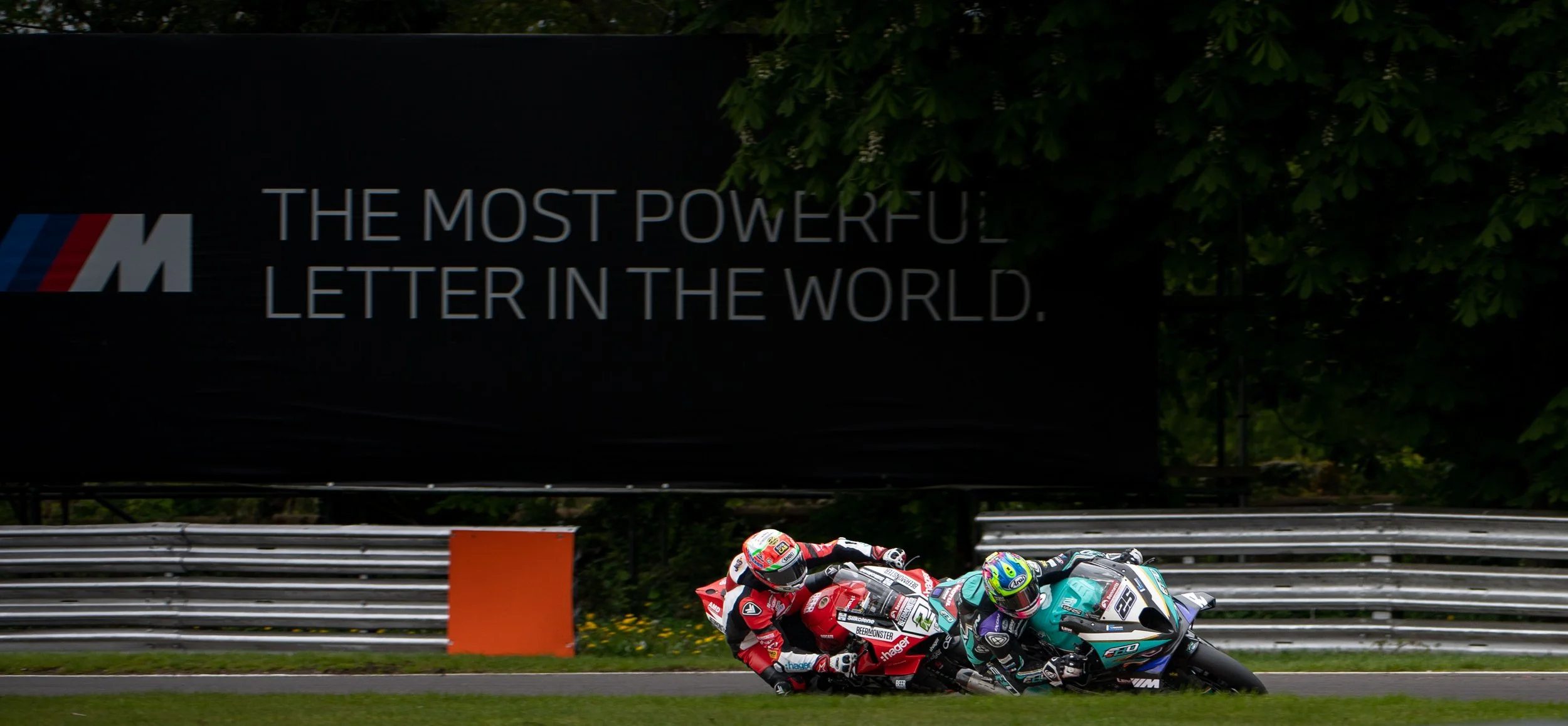 Two motorcycle racers leaning into a turn on a racetrack, with a black advertisement banner reading 'THE MOST POWERFUL LETTER IN THE WORLD' and a BMW M logo in the background.