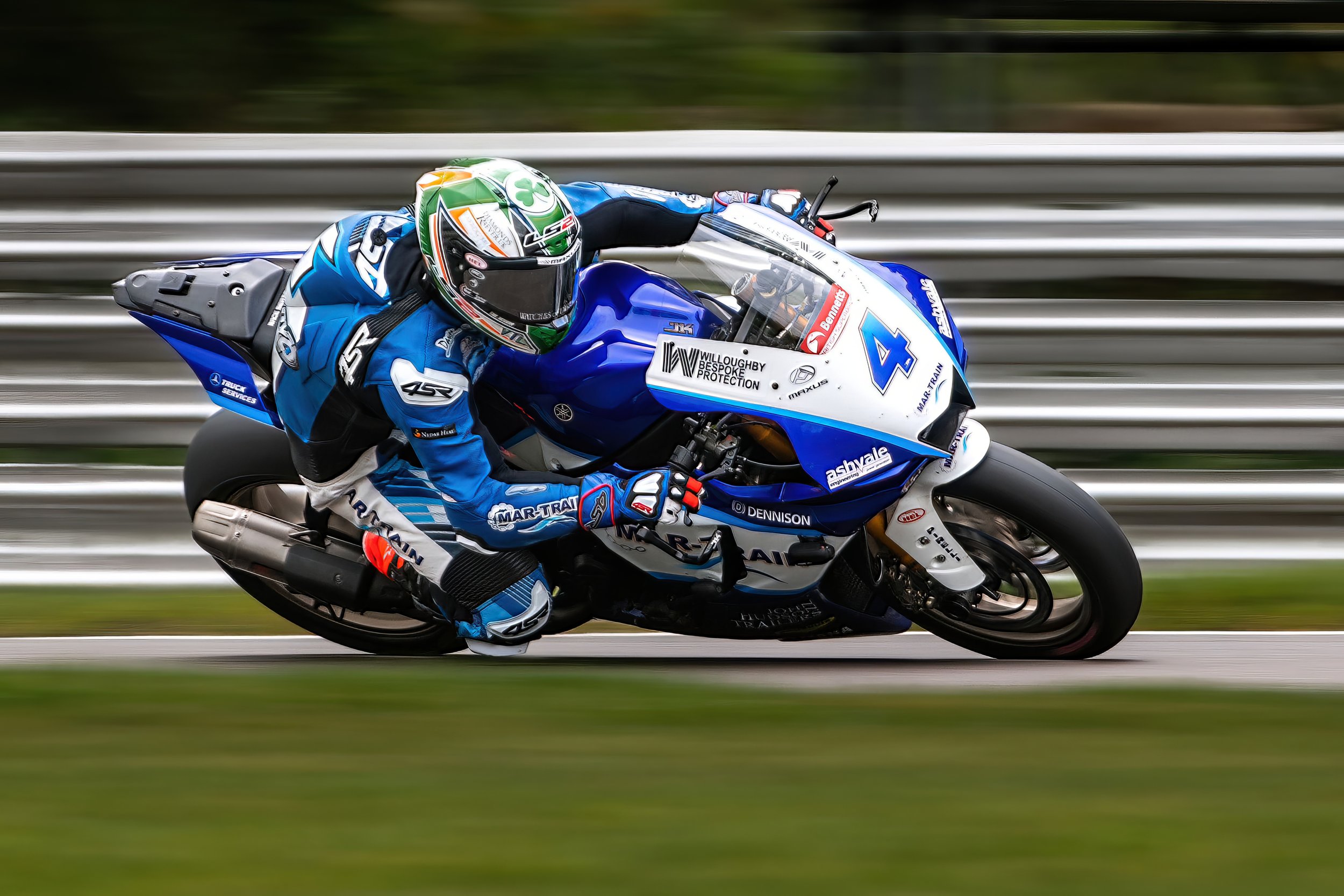 Motorcycle racer dressed in blue racing suit, helmet, and gloves leaning into a turn on a race track with striped barriers in the background.