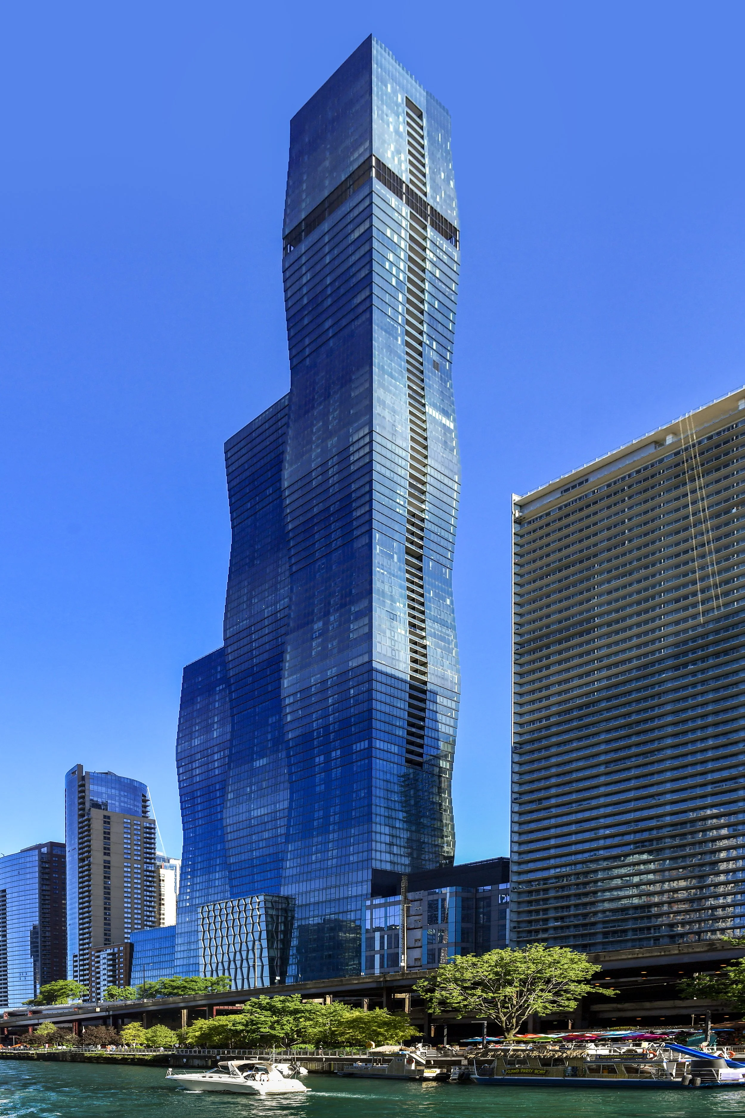 Tall, modern glass skyscraper with a twisted, irregular design in an urban waterfront area, surrounded by other high-rise buildings, trees, and boats on the water under a clear blue sky.