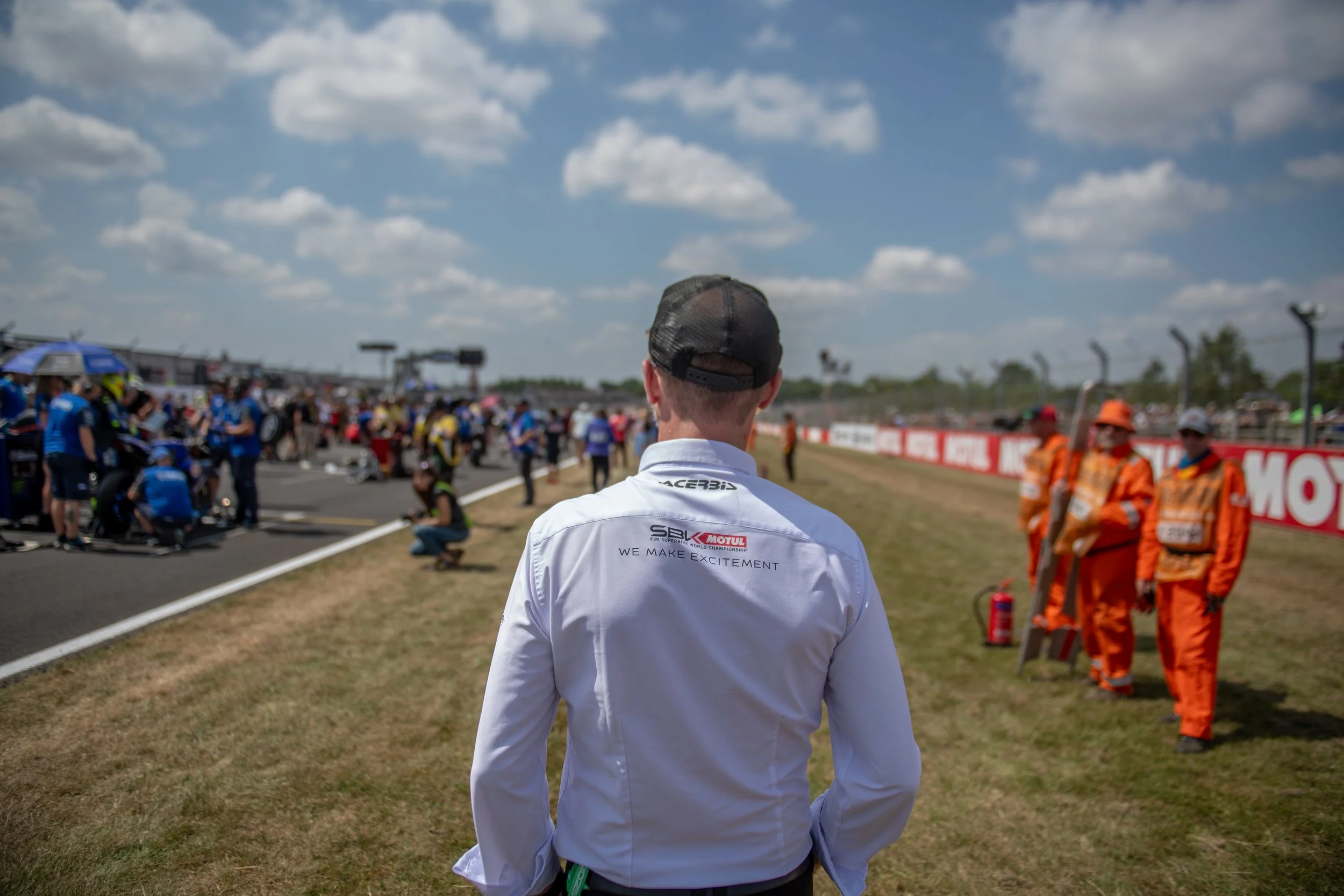 Person wearing a white shirt with sponsor logos, observing a busy race track scene with team members, officials, and spectators on a sunny day.