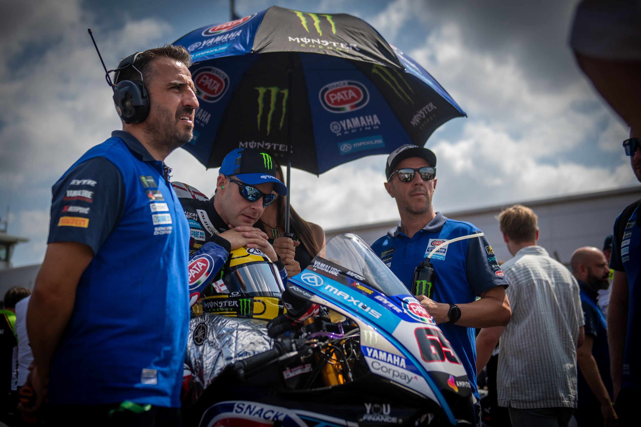 A group of men in blue team shirts gathered around a Yamaha racing motorcycle, with one man wearing sunglasses and holding a large umbrella with various sponsor logos, including Monster Energy and Yamaha. One person appears to be the rider, sitting o