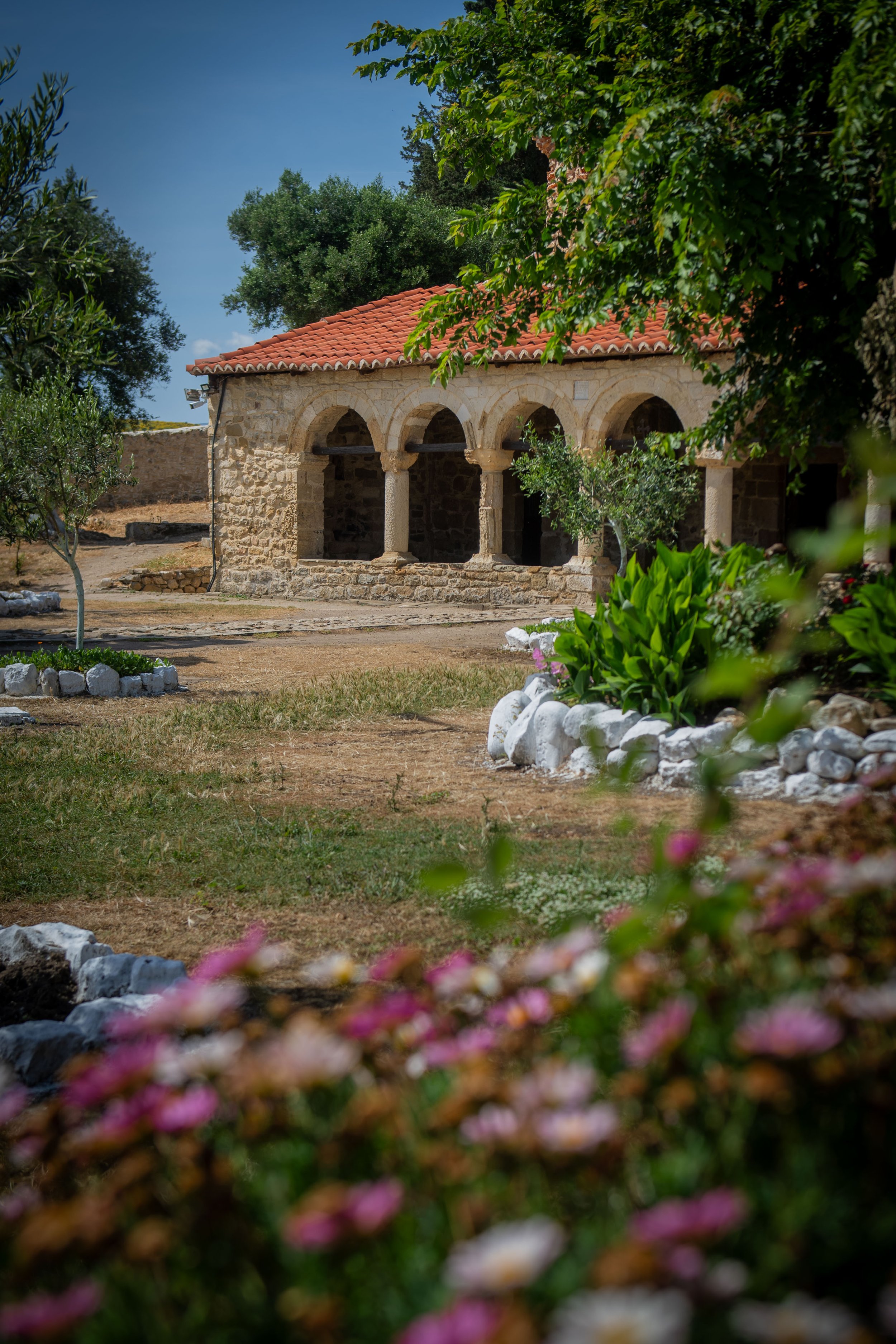 A stone building with a red tile roof, arched openings, and stone columns, surrounded by green trees, bushes, and flowers under a blue sky.