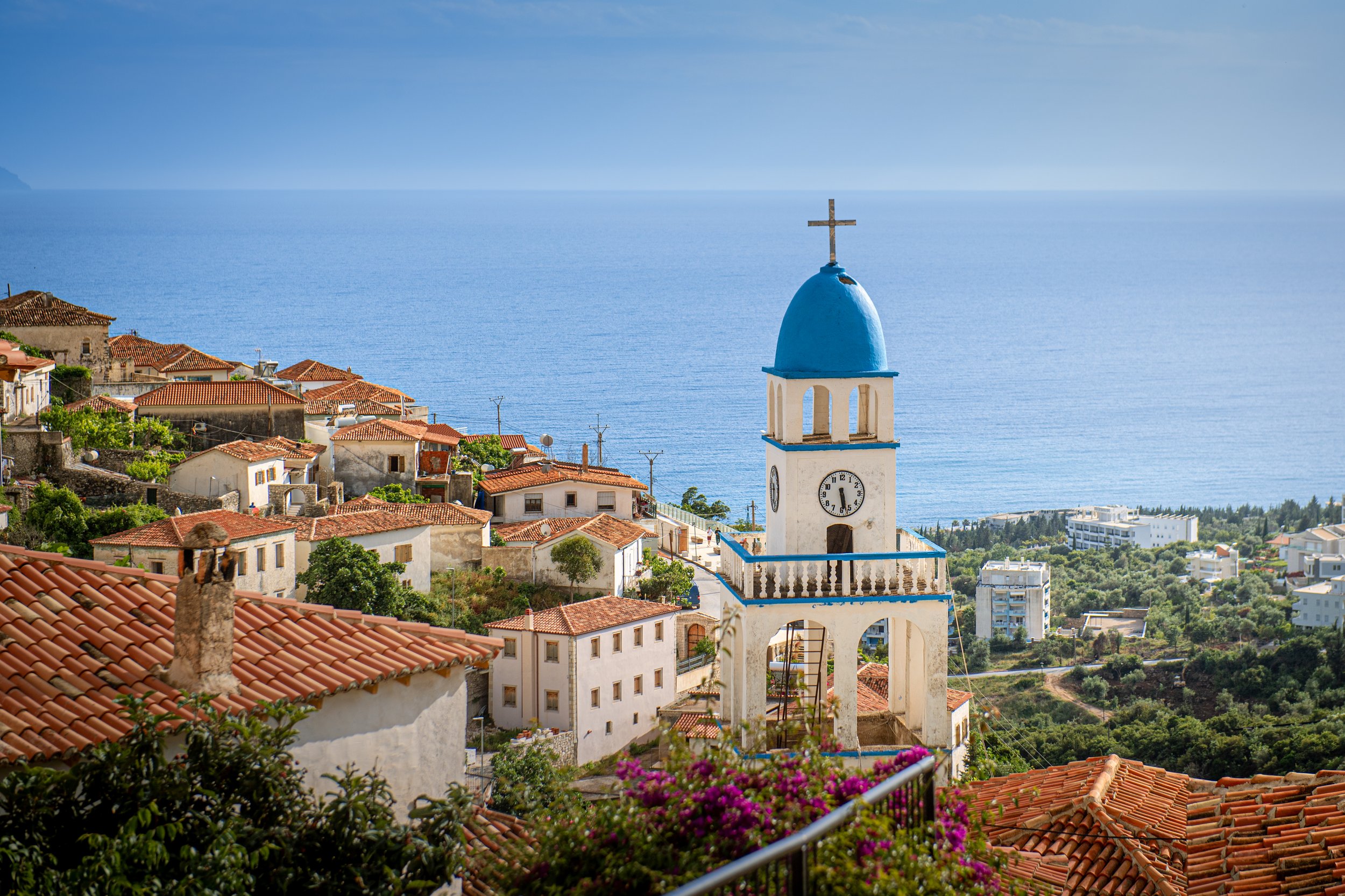 A coastal village with white buildings and red-tiled roofs, a white church with a blue dome and cross, overlooking the ocean on a sunny day.