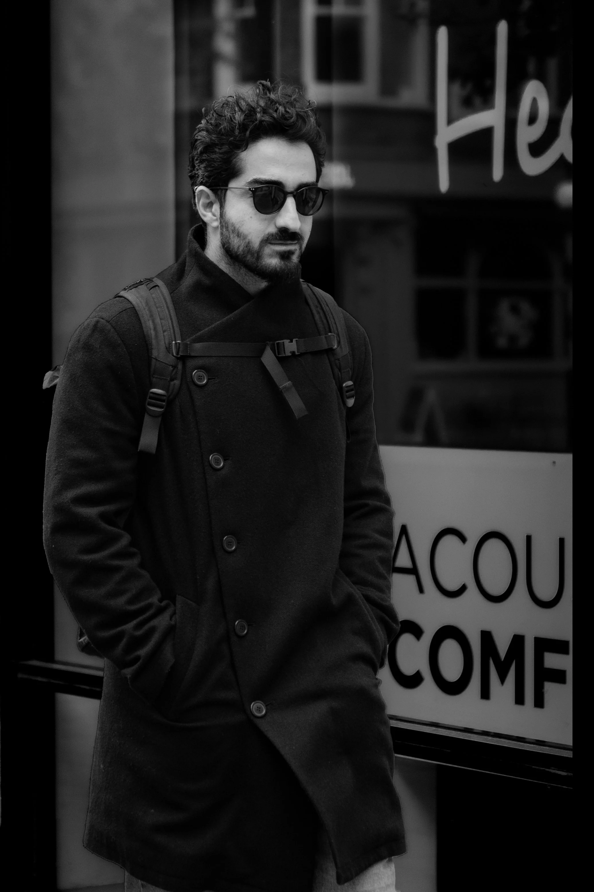 A man with curly hair, beard, and sunglasses walking on the street, wearing a dark coat and backpack, in front of a storefront window with signs.