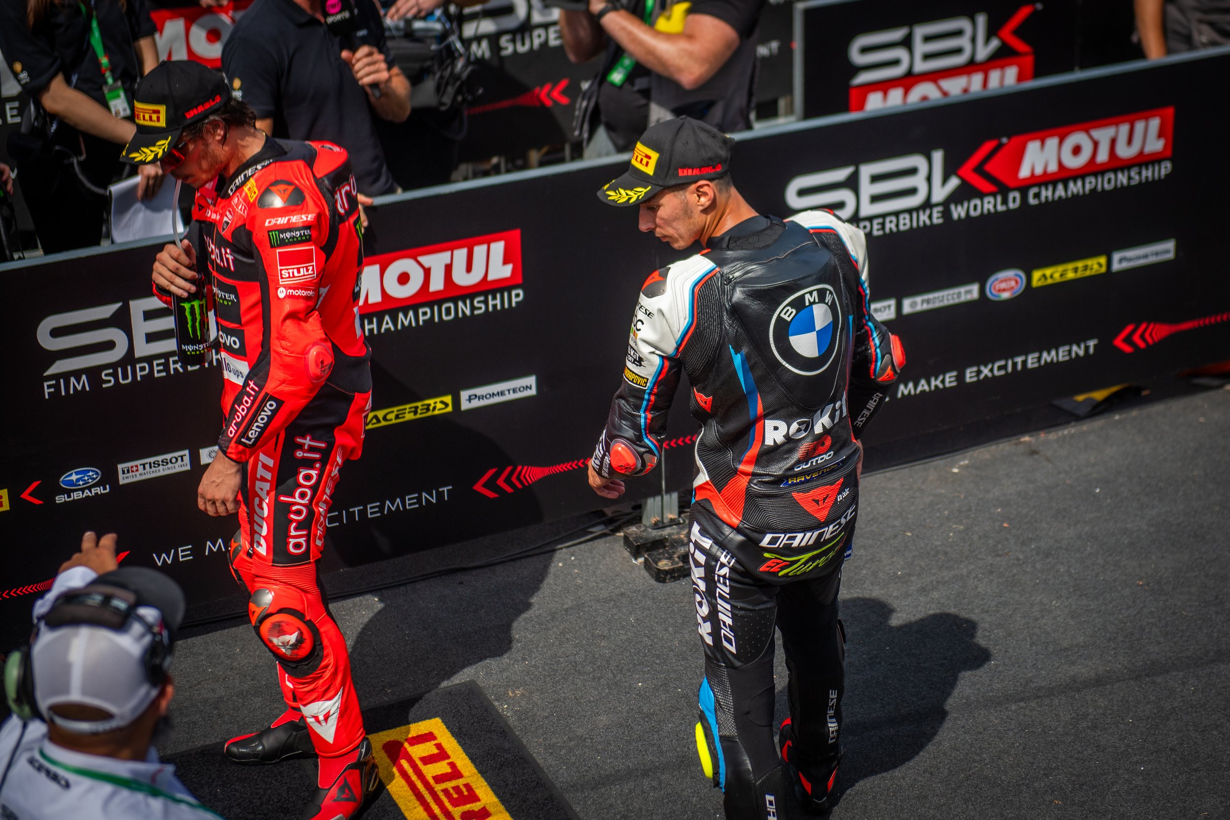 Two motorcycle racers in racing suits and caps preparing for a race, standing behind a barrier with sponsors' logos, including Motul, Subaru, and BMW, at a Superbike World Championship event.