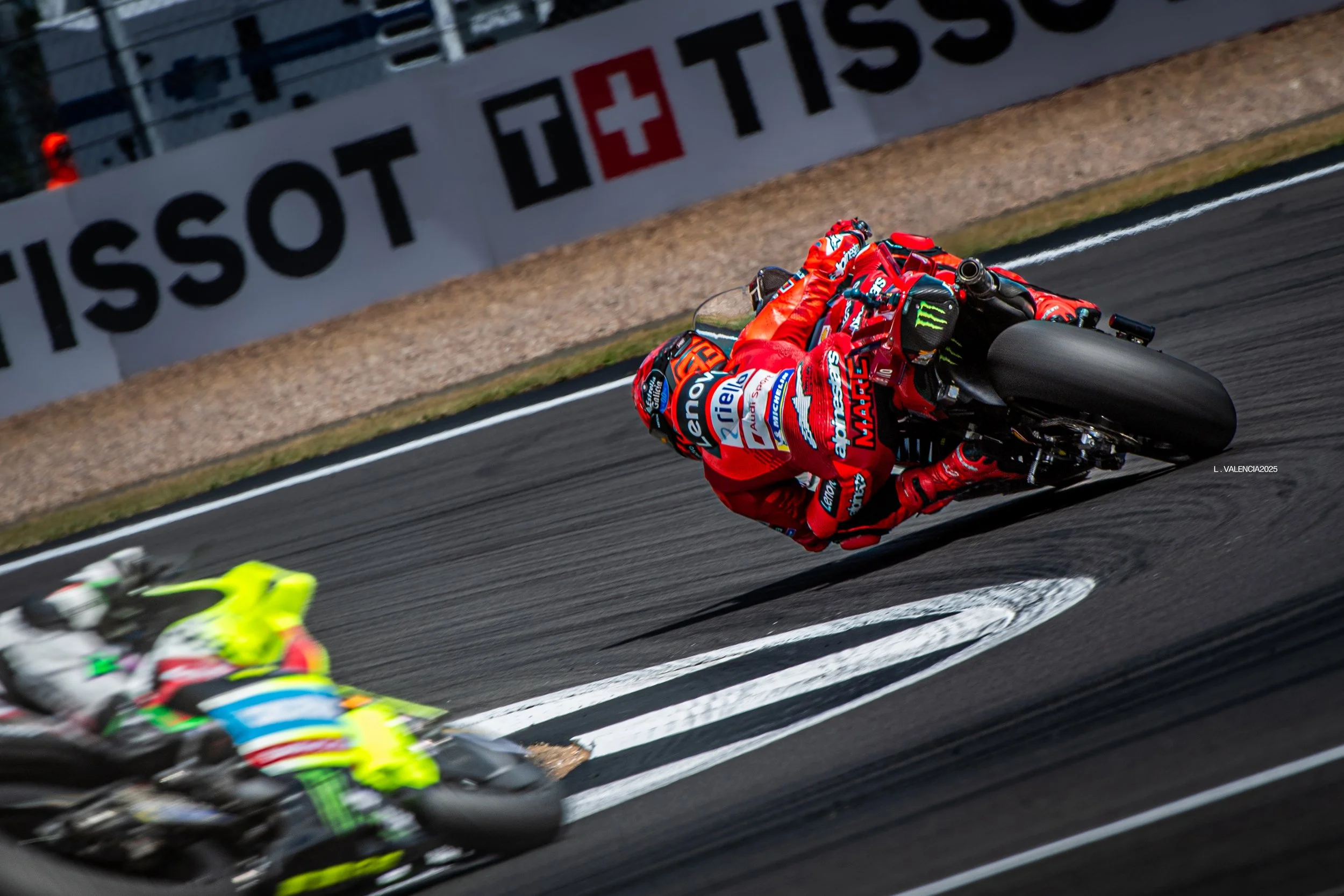 Two motorcycle racers leaning into a sharp turn on a racing track, one in bright yellow and green gear, the other in red gear, with branding and sponsor logos visible on their suits and bikes, and a Tissot advertisement in the background.