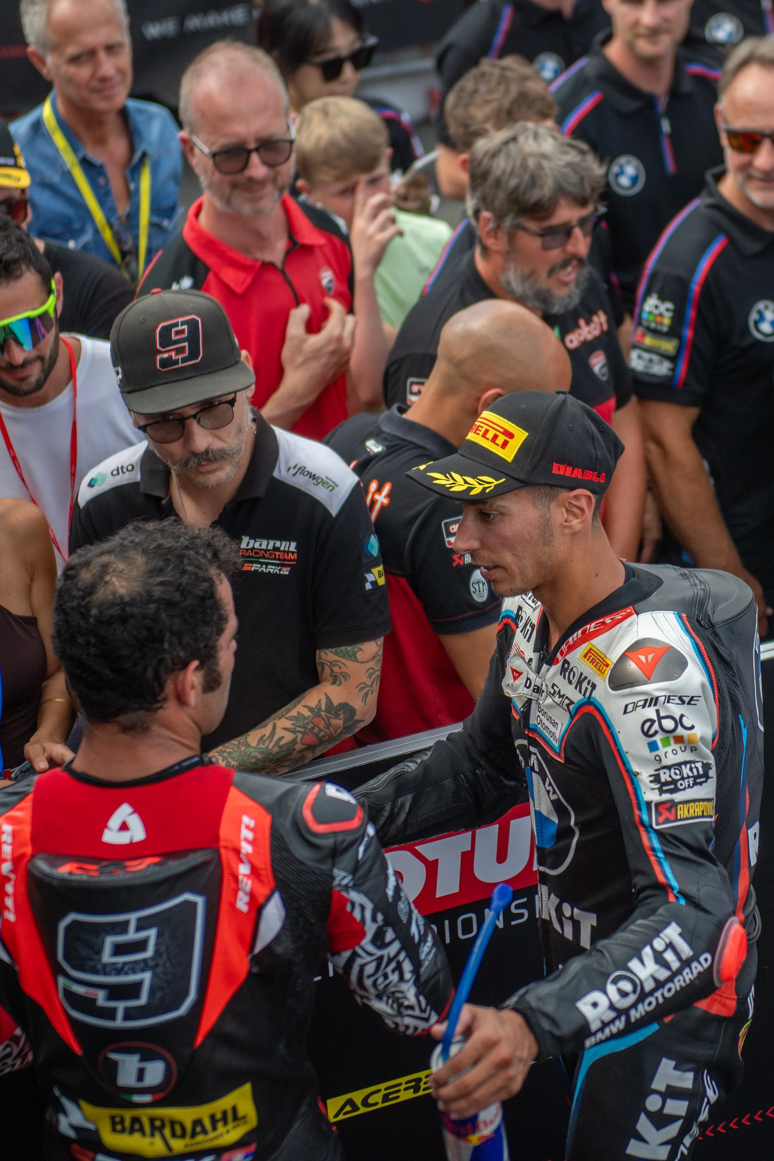 Motorcycle racers in racing suits and caps greeting each other, with a crowd of spectators behind them at a motorsport event.