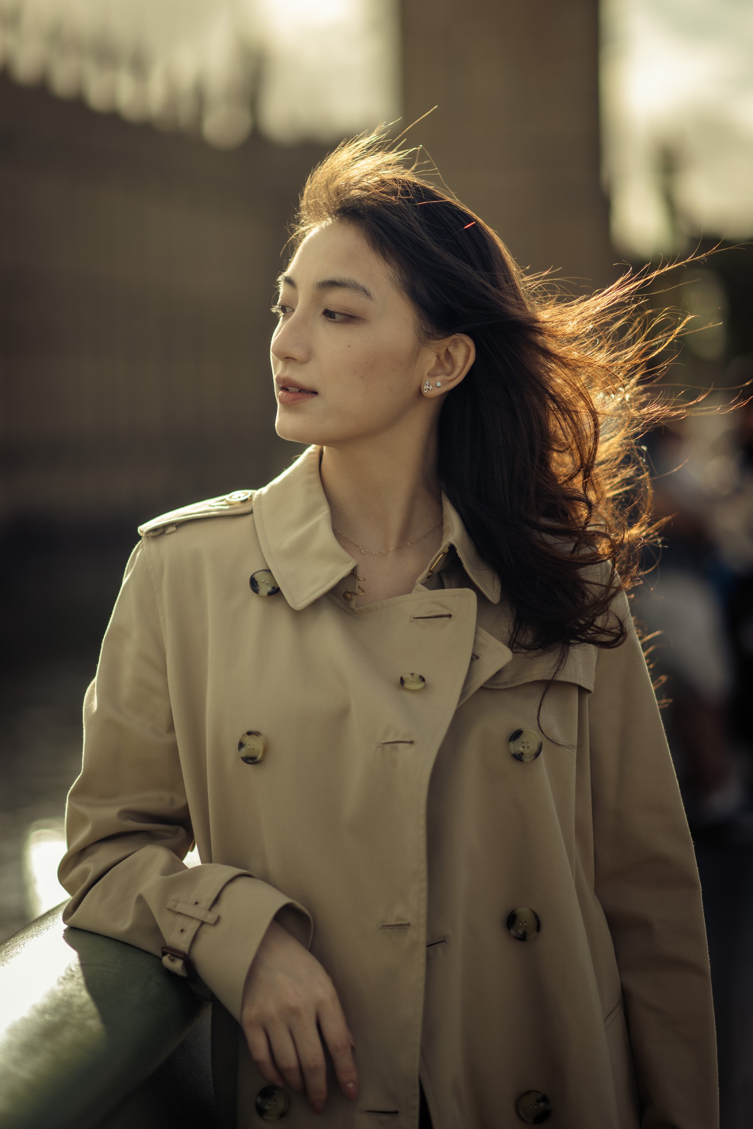 A woman with long dark hair wearing a beige trench coat leaning on a railing outdoors.