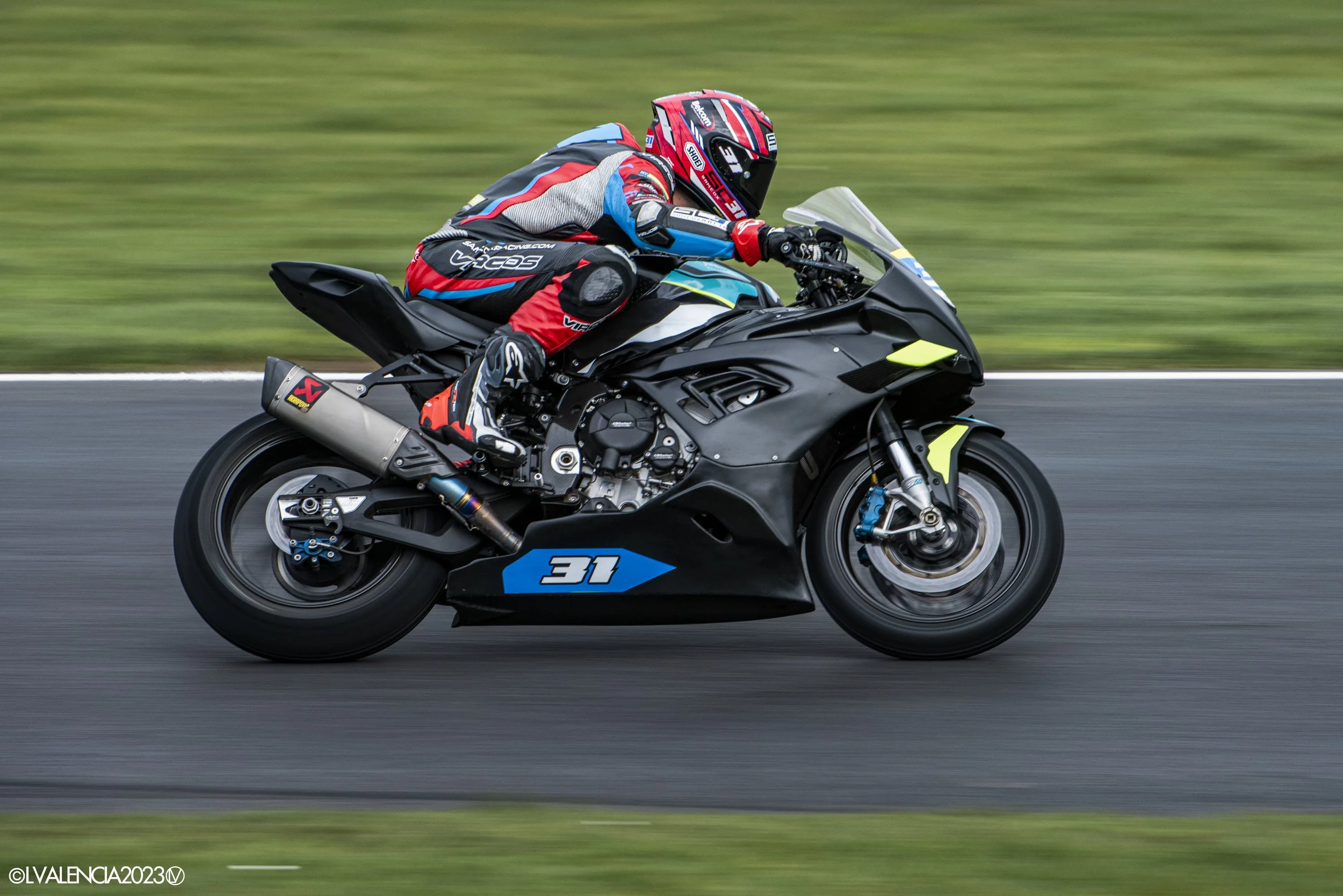 A motorcycle rider leaning into a turn on a racetrack, wearing a red, black, and blue racing suit and helmet, on a black racing motorcycle with the number 31.