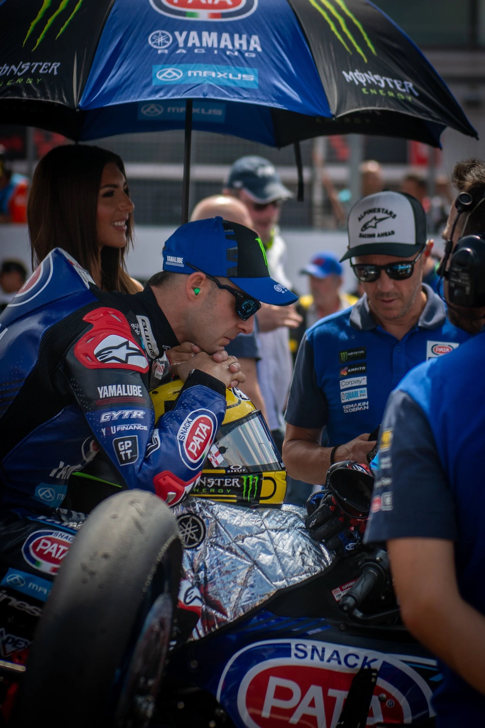 A professional motorcycle racer in racing gear, holding his helmet with a PATA snack logo, is deep in thought under a blue and black umbrella with sponsorship logos. Several team members and people are around him, some wearing sunglasses and team uni