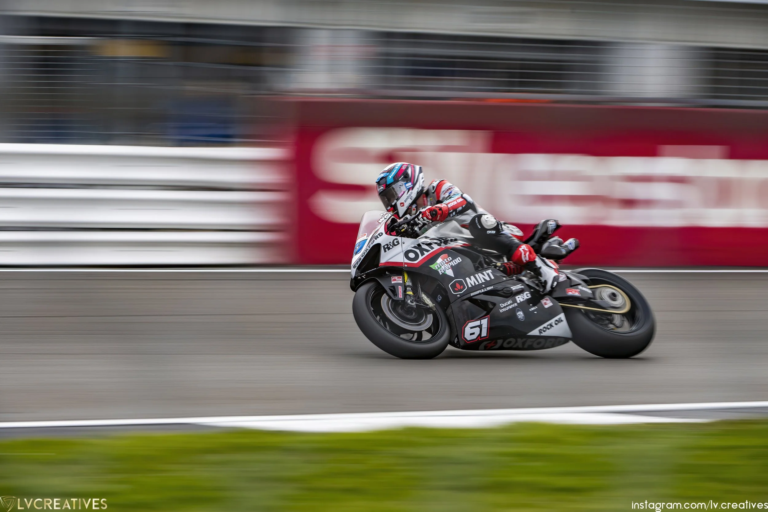 Motorcycle racer in black and white racing suit leaning into a turn on a black motorcycle with the number 61, on a race track with blurred background.