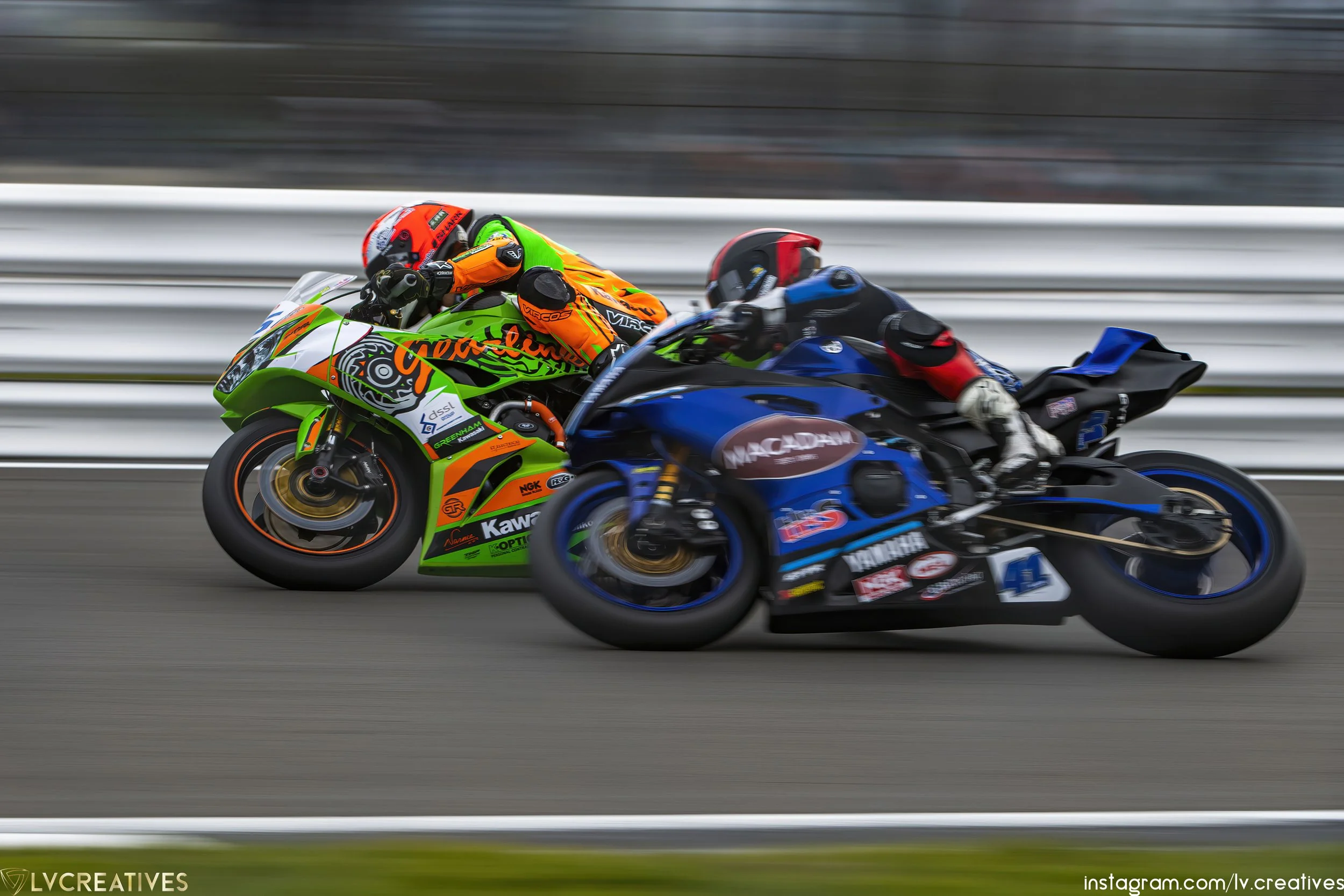 Two motorcycle racers leaning into a turn on the racetrack, one in a bright green and orange suit with a matching green and orange motorcycle, the other in a black suit with a blue motorcycle, both motorcycles in motion with blurred background.