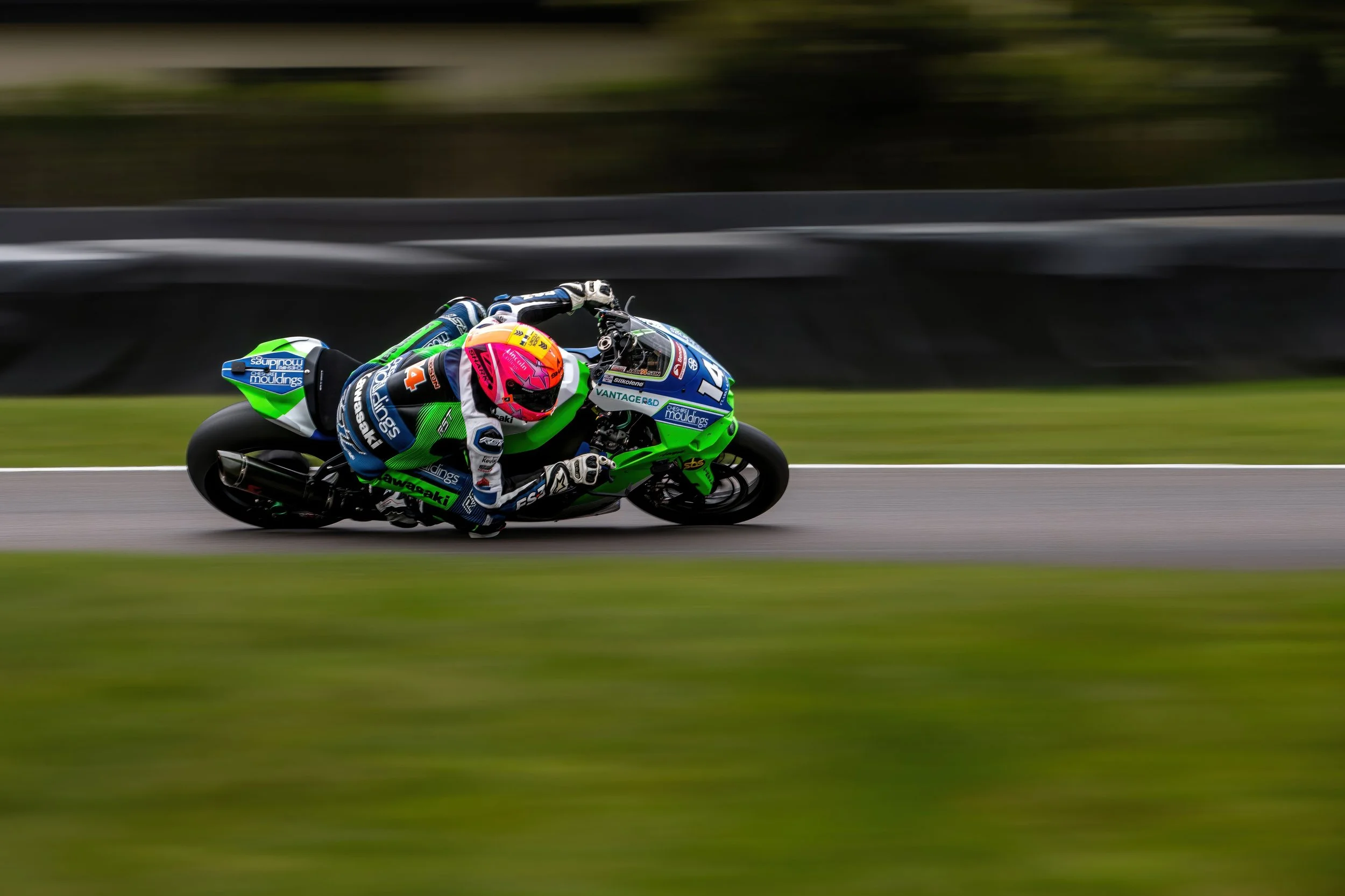 Motorcycle racer in a green and blue suit and pink helmet leaning into a turn on a racetrack.