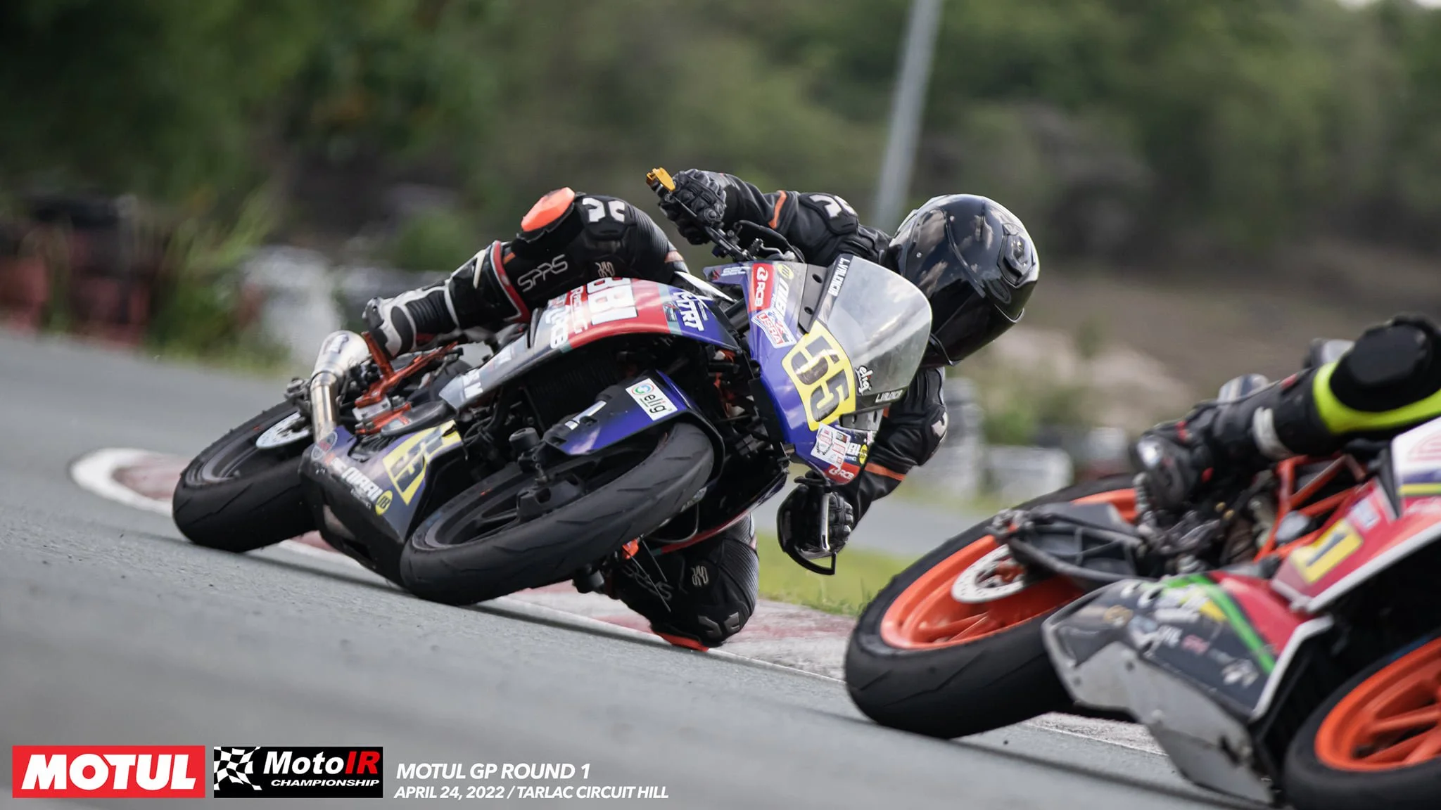 Motorcycle racers leaning into a turn during a race on a curvy road, with lush green trees in the background.Lawrence Valencia and Carl Ferrer's intense battle