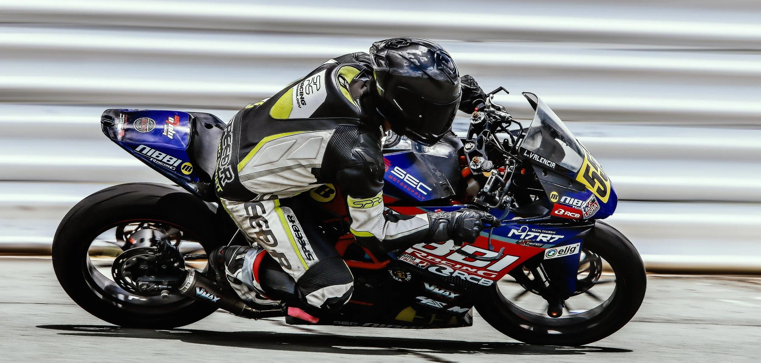 Lawrence Valencia wearing black helmet and racing suit leaning into a turn on a race track, with a blue, red, and yellow motorcycle marked with various sponsor logos.