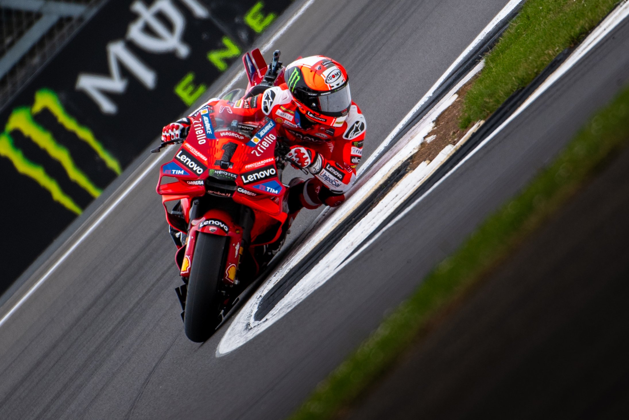 A professional motorcycle racer in a racing suit and helmet riding a red Ducati motorcycle on a race track with branding and advertisements in the background.