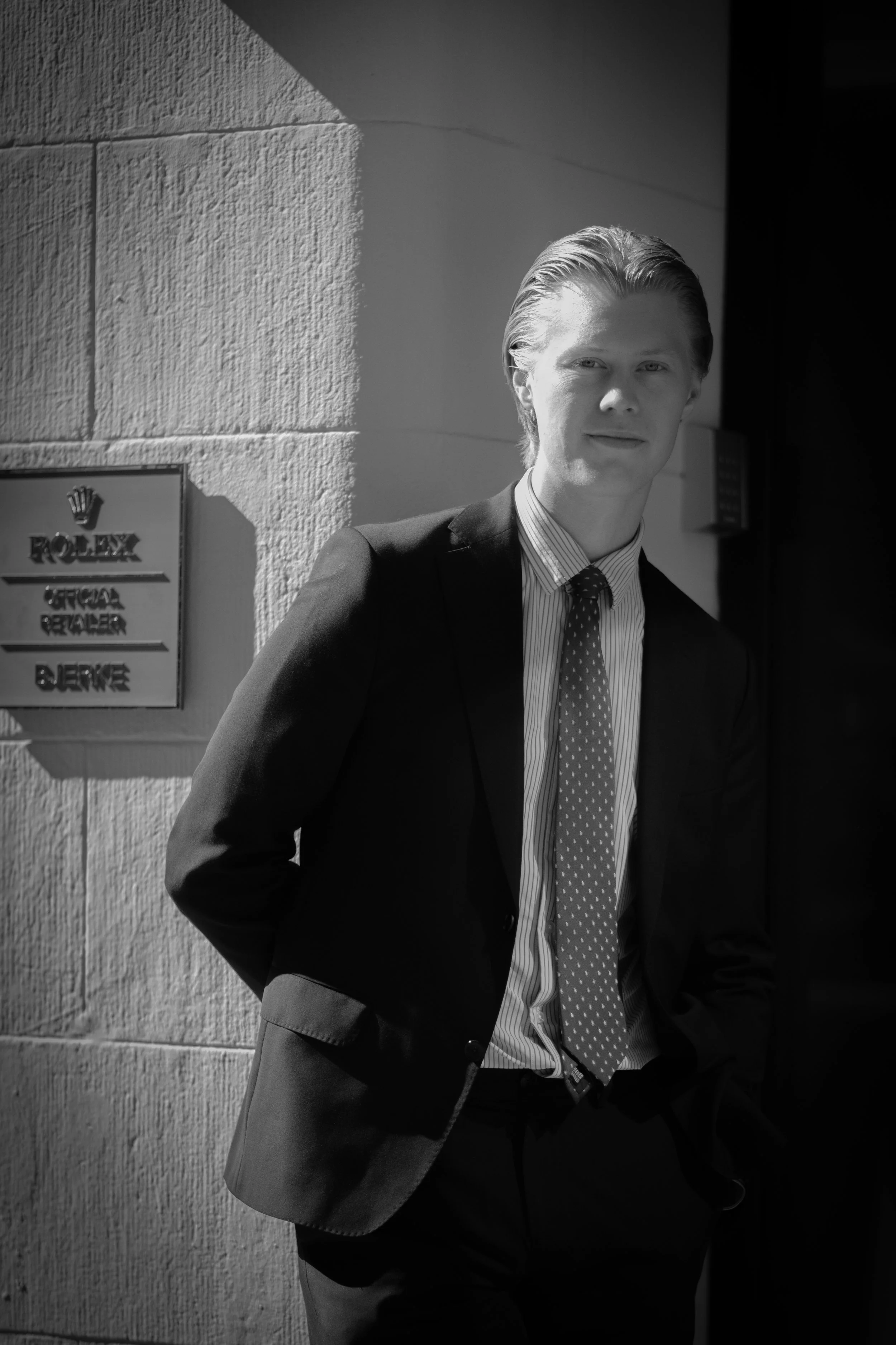 A young man in a suit and tie leaning against a textured wall with a Rolex sign on it.