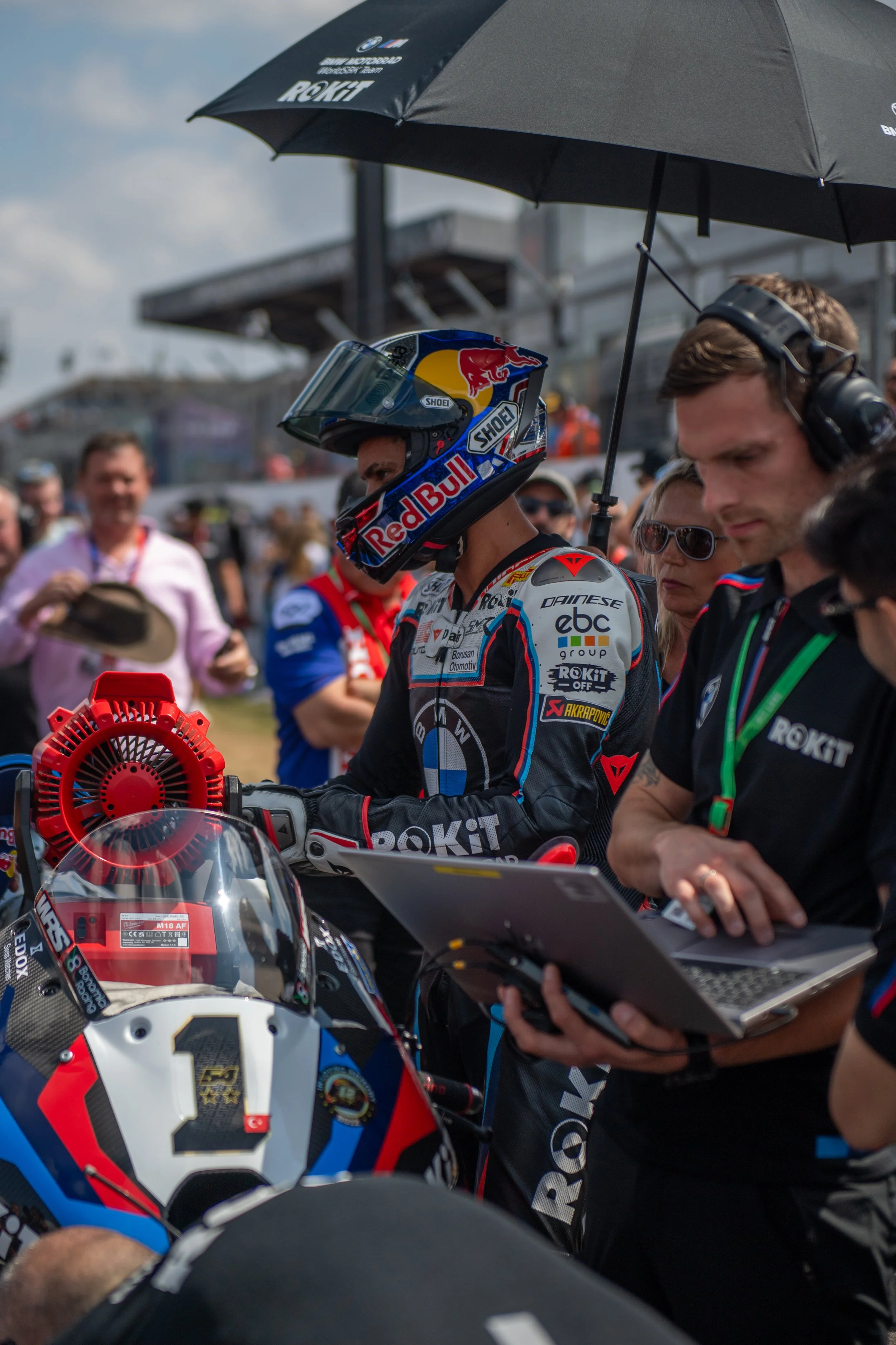 A motorcycle racer in a helmet and racing gear standing in a team area with team members, holding a laptop, under an umbrella.