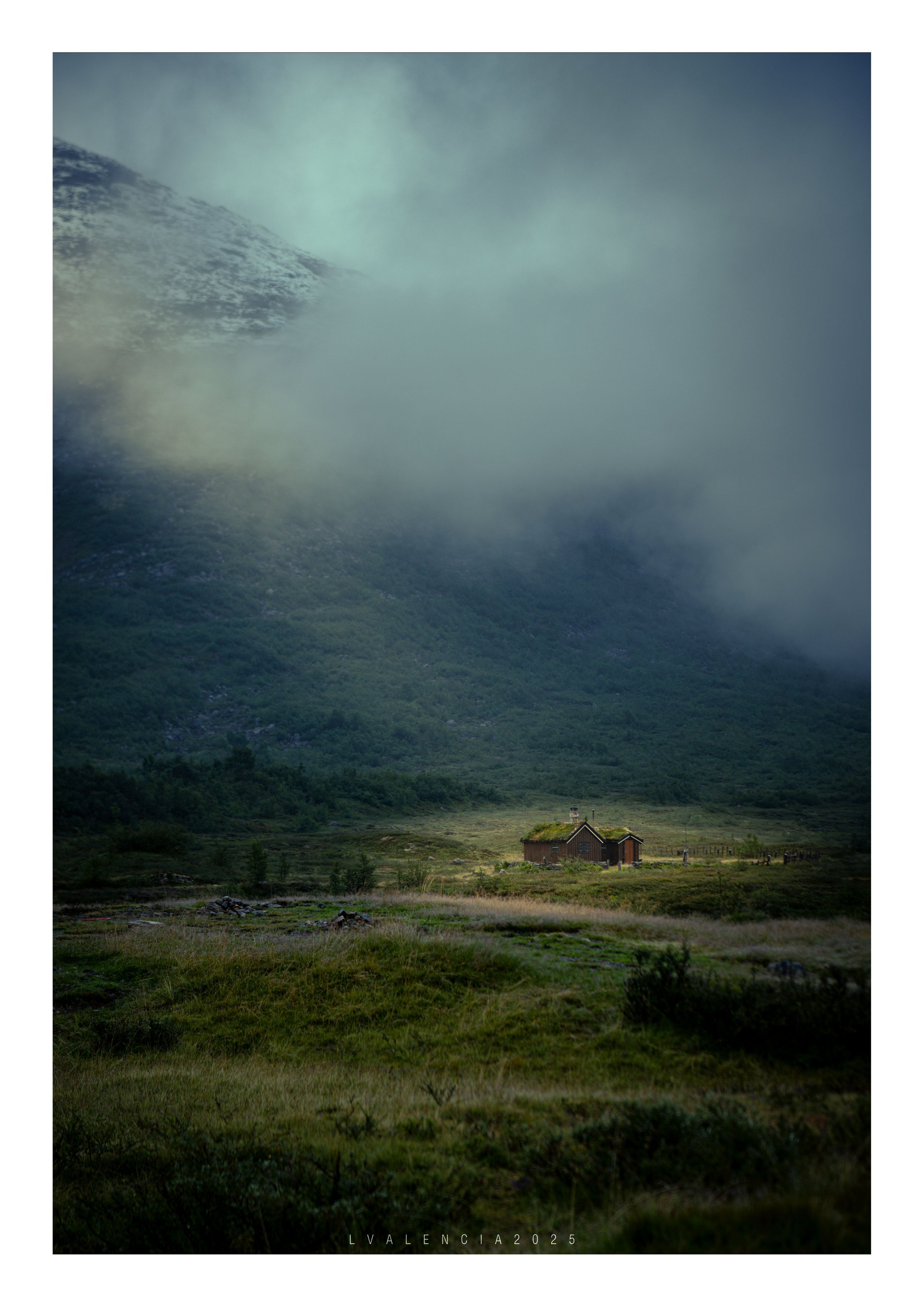 A small house with a green roof sits in a vast green valley with mountains and clouds in the background.