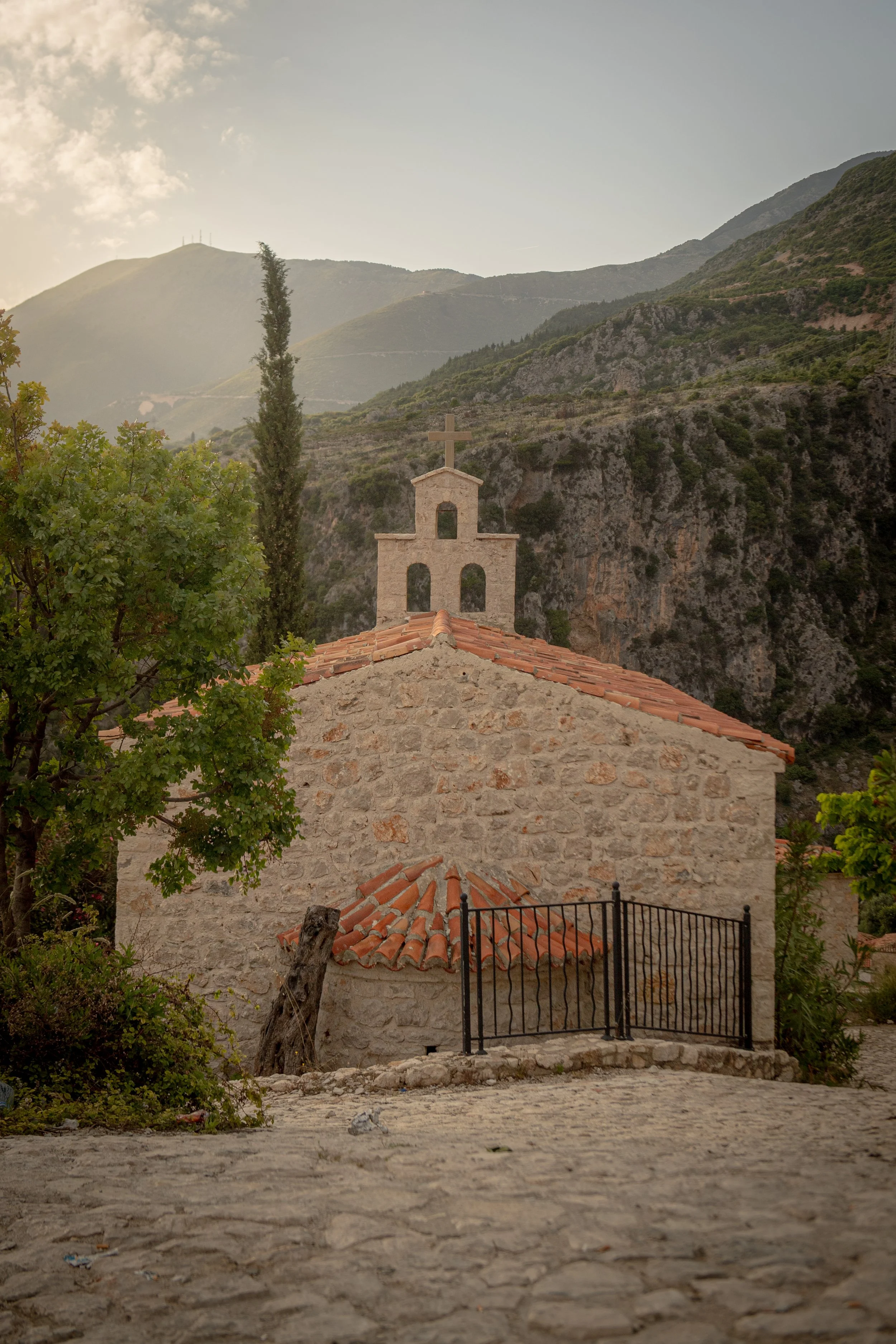 A small stone church with a red-tile roof and a cross on its steeple, surrounded by greenery and mountains in the background, under a cloudy sky.