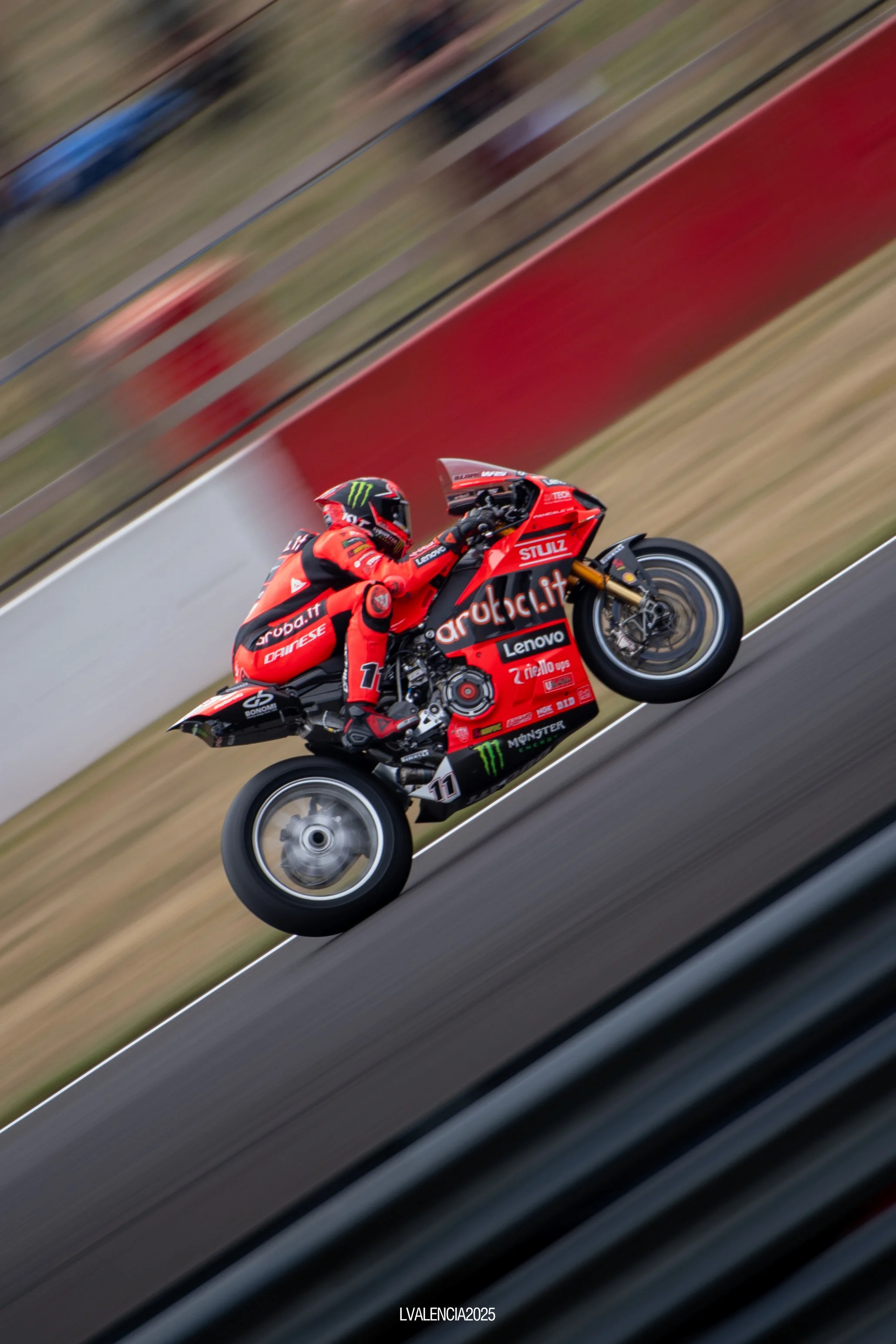 A motorcycle racer in red gear and helmet performing a wheelie on a red MotoGP bike on a race track.