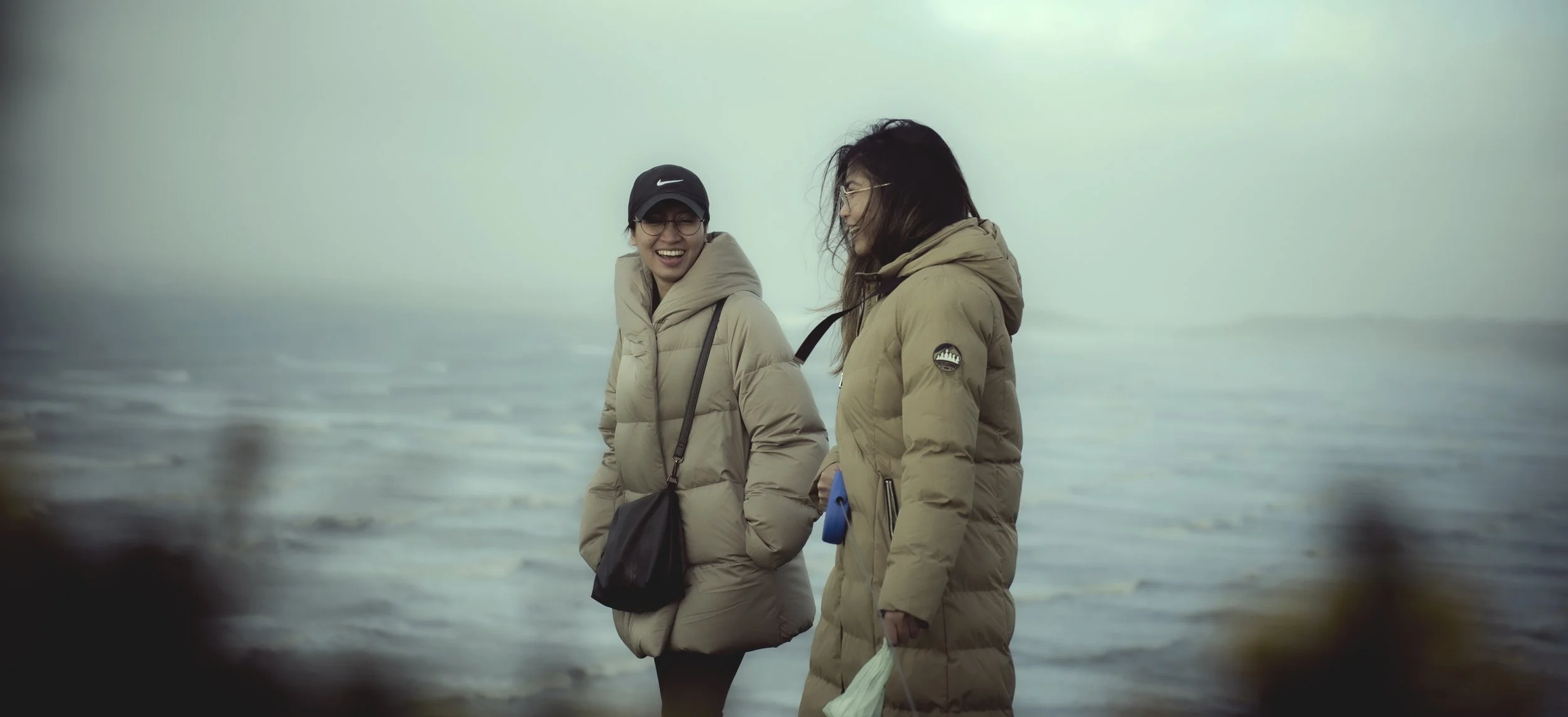 Two women in beige winter coats walking on the beach, smiling and talking, with ocean waves in the background.