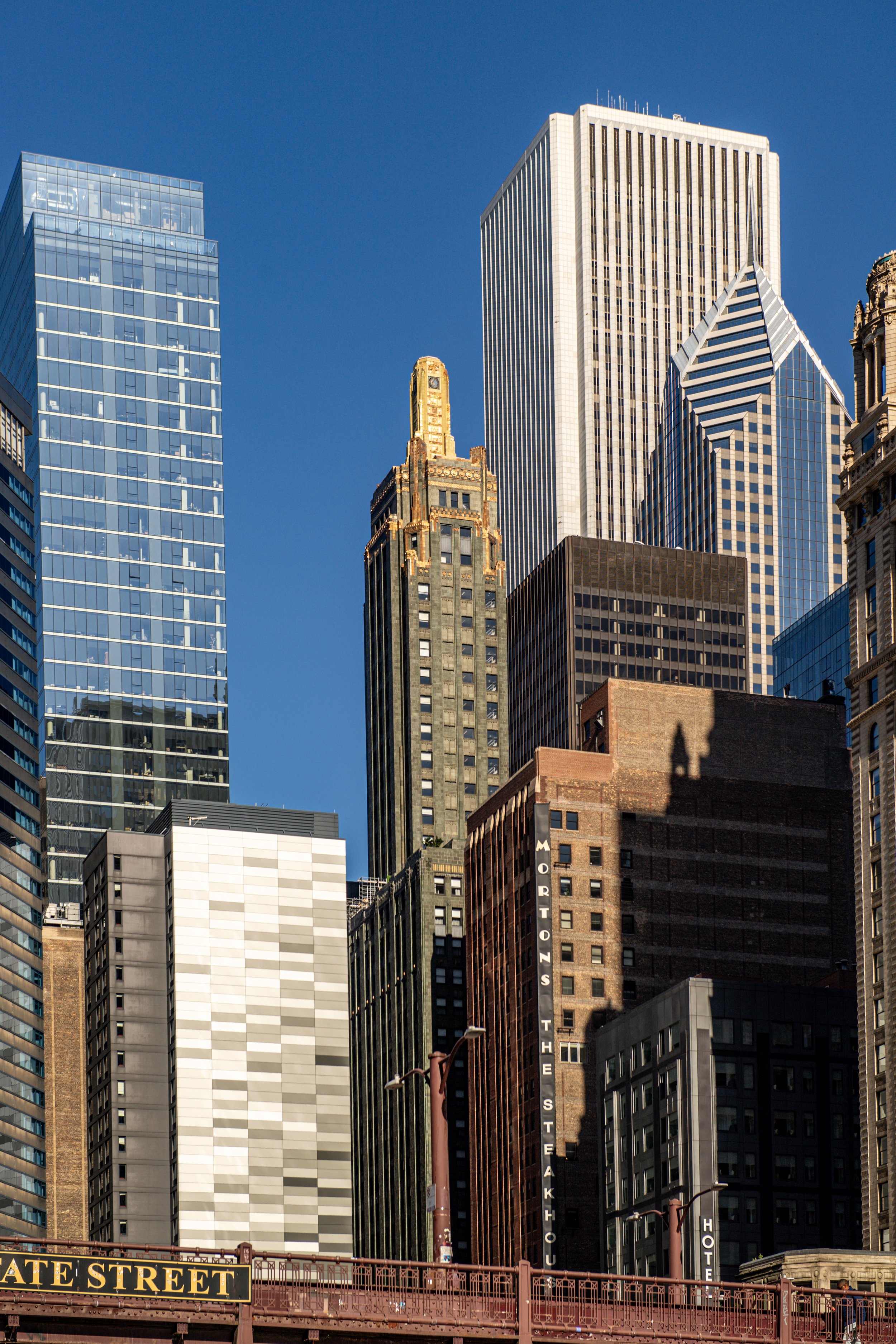 A cityscape featuring various tall skyscrapers with glass and stone facades against a clear blue sky. The bottom shows a bridge with a sign reading 'State Street'.