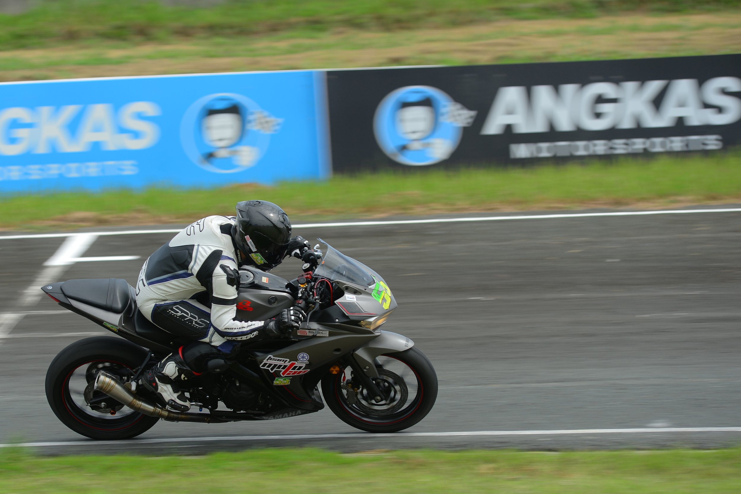 Motorcycle racer in black and white racing suit on a black Yamaha bike, speeding on a racetrack with a blue and black advertising banner in the background.