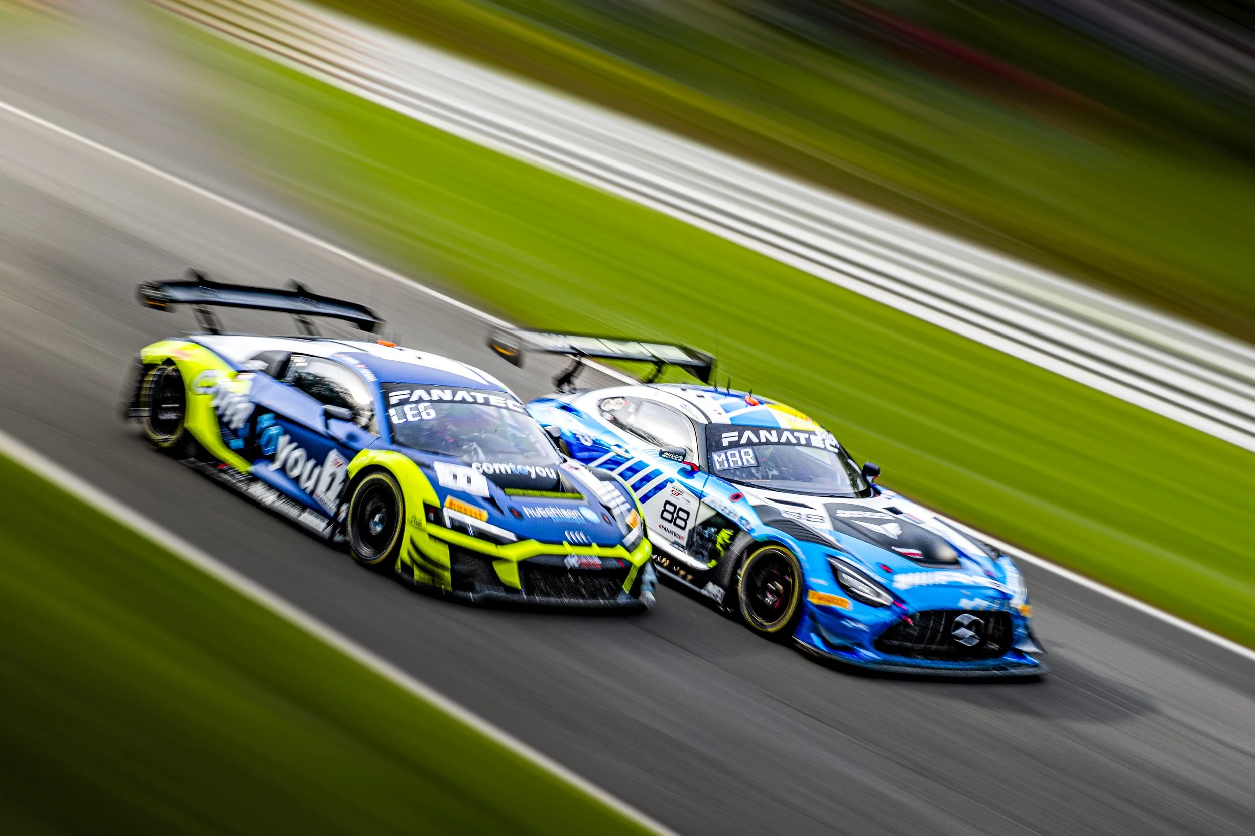 Two race cars speeding on a race track, with motion blur and a green grassy background.