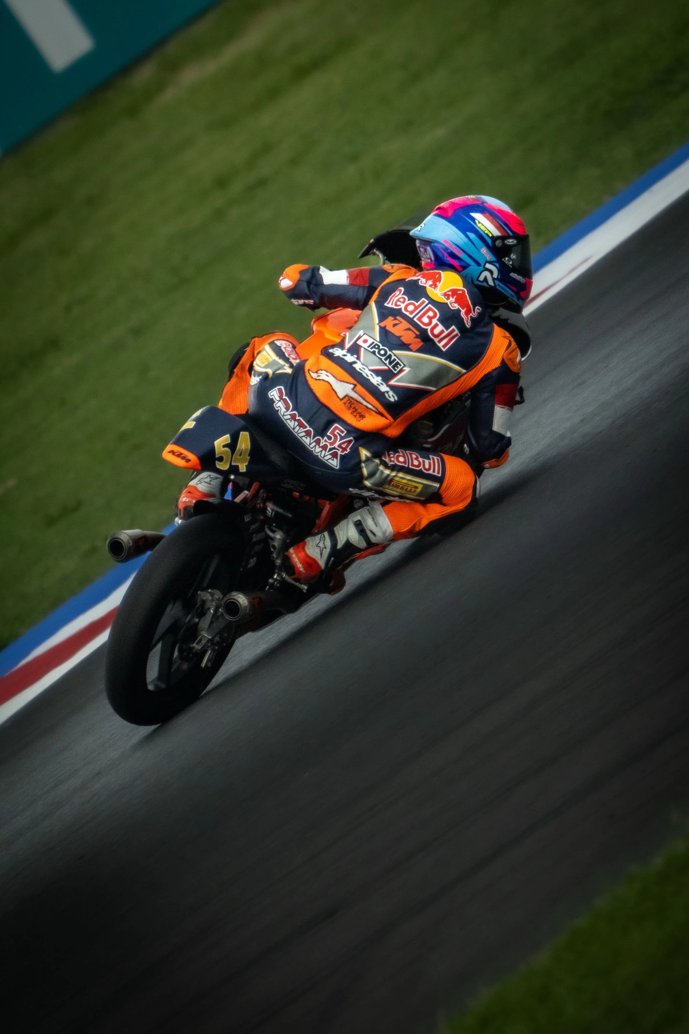 A motorcycle racer in orange and black racing gear, with a blue and pink helmet, leaning into a curve on a racetrack. The bike has the number 54 and sponsors like Red Bull and KTM.