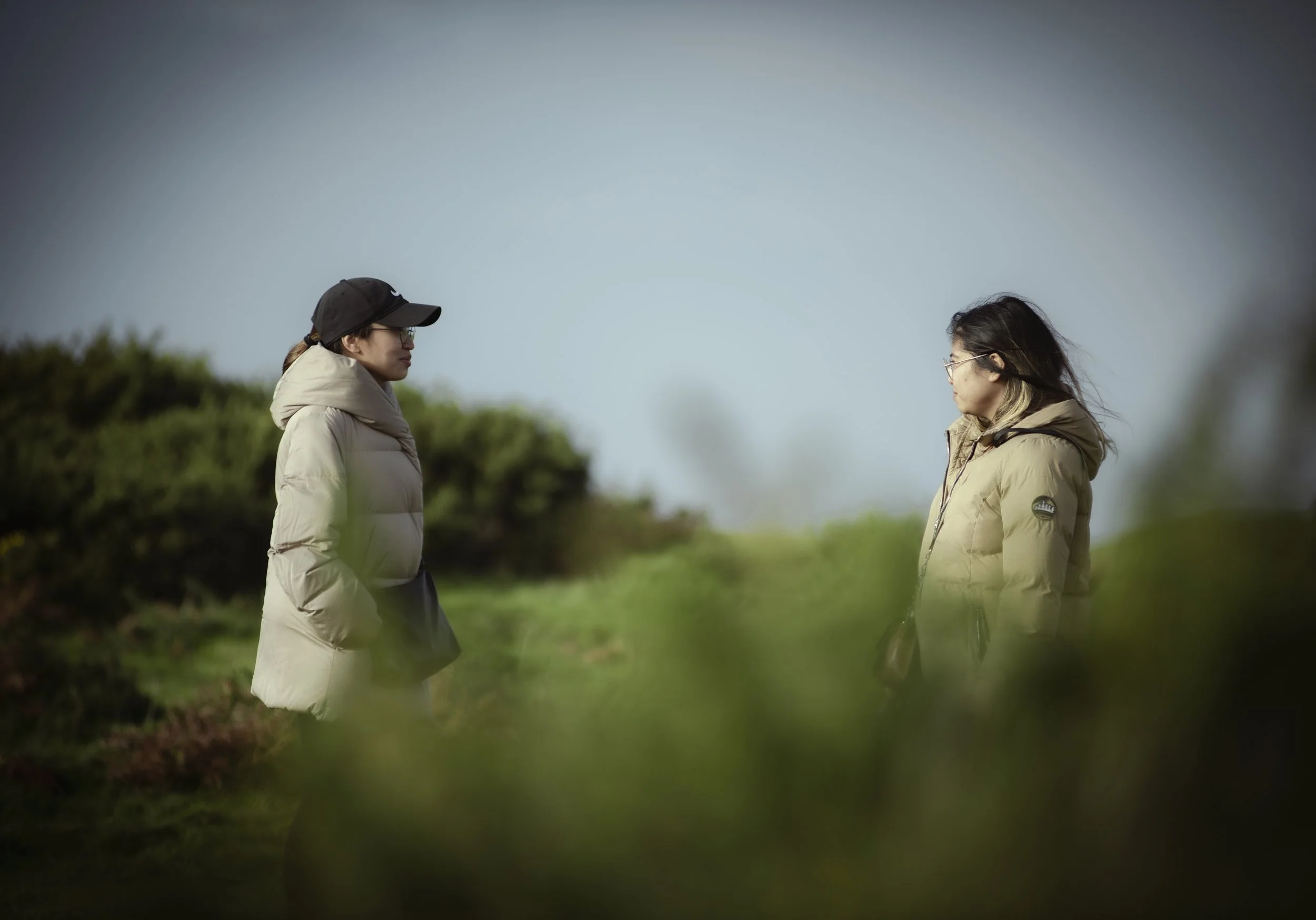 Two women in beige jackets facing each other outdoors on a cloudy day, surrounded by greenery.